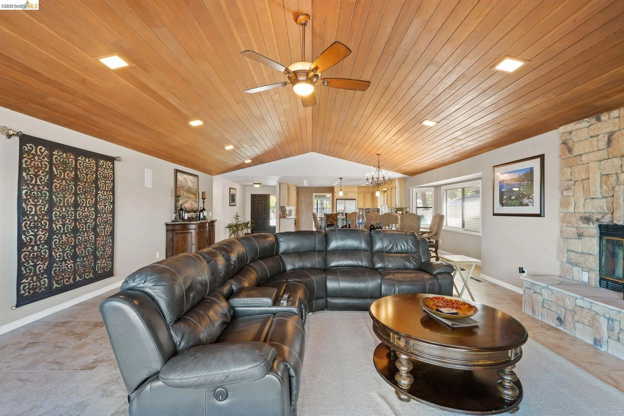 Living room with leather sectional sofa, wooden coffee table, stone fireplace, large windows, and wood-paneled ceiling with ceiling fan.