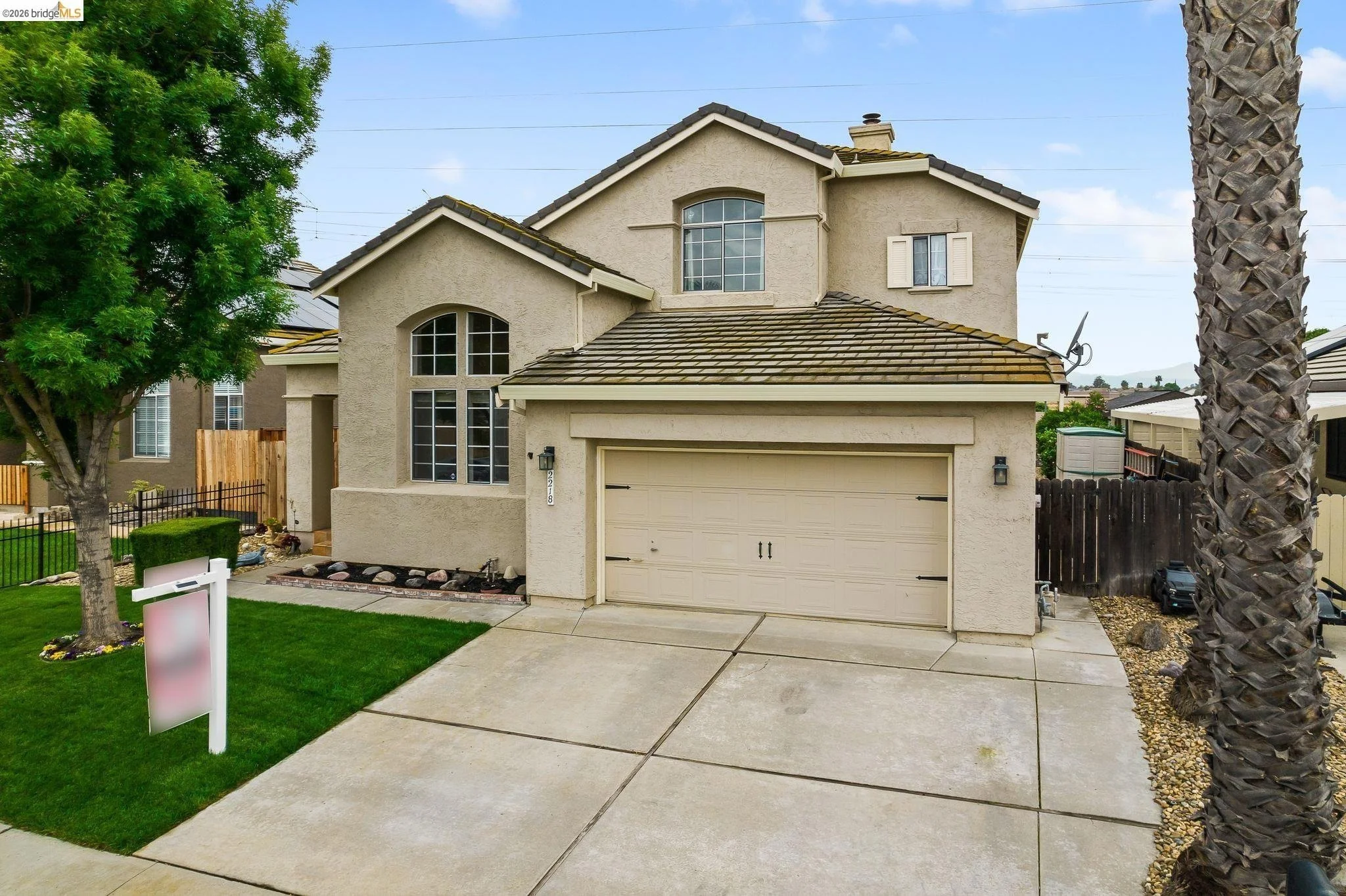 Two-story beige house with a brown tiled roof, large arched windows, and a front lawn with a tree and a white sign. The driveway leads to a closed garage door.