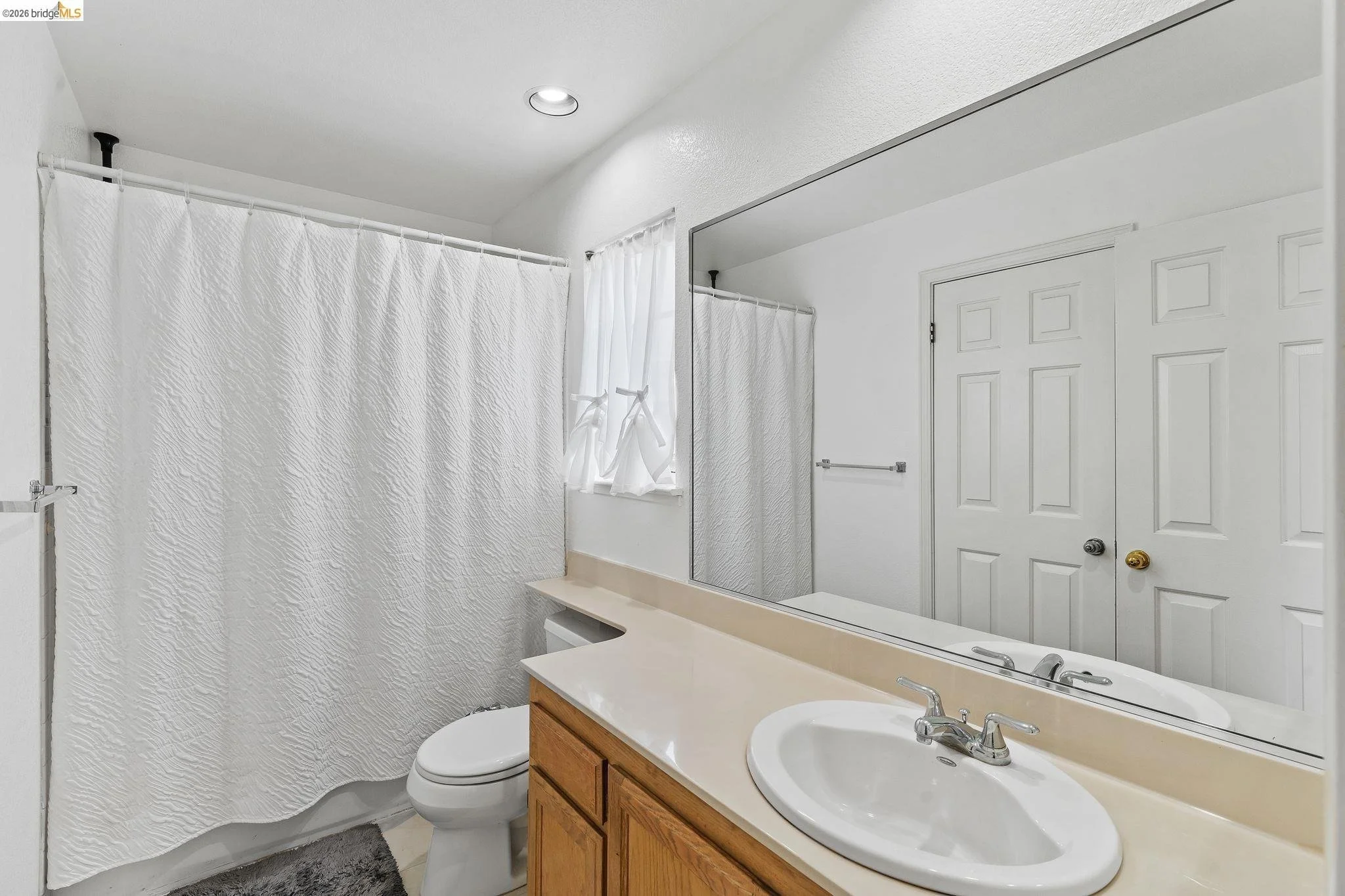Bathroom with white walls, a large mirror, a single sink vanity with wooden cabinetry, a toilet, and a shower with a white textured curtain. There is a small window with white curtains and a towel bar on the wall.