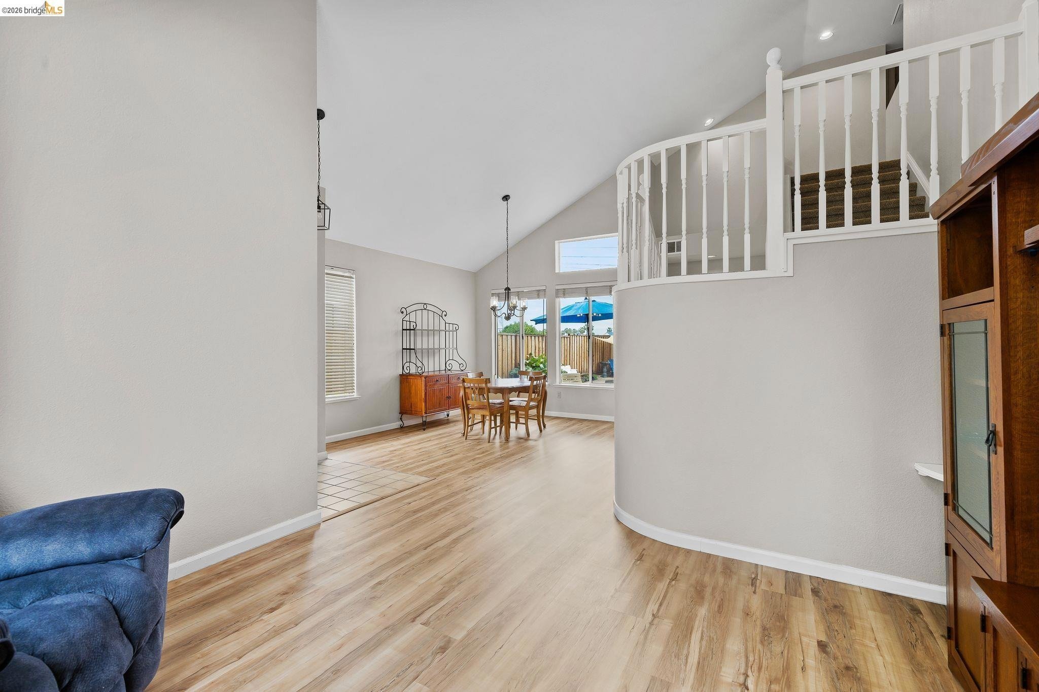 Interior view of a living and dining area with hardwood flooring, white walls, a curved staircase with white balusters, a dining table with chairs, large windows with blue umbrellas outside, and a wooden hutch.