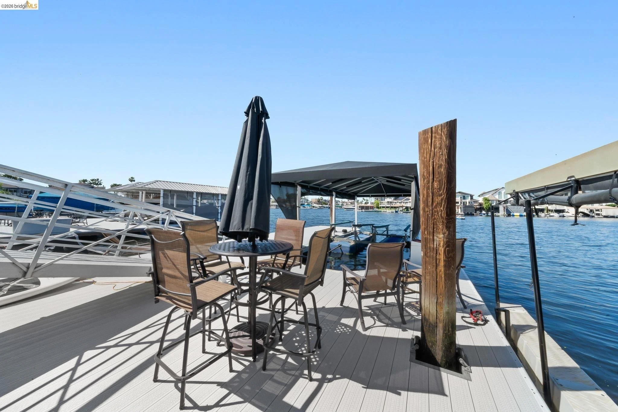Outdoor patio on a boat dock with a round table, six chairs, a black umbrella, a boat covered with a canopy, and water with houses in the background during daytime.
