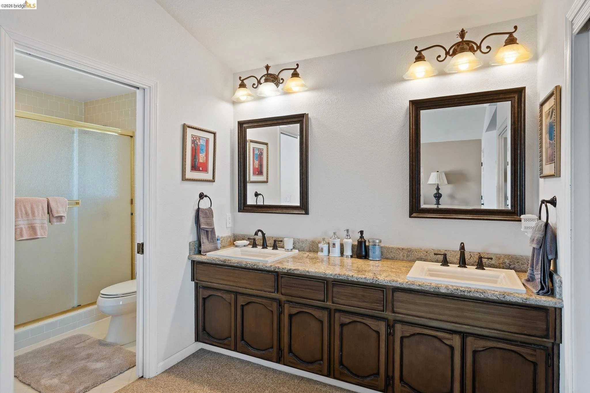 Bathroom with a double vanity sink, granite countertop, and dark wood cabinets. Two mirrors above each sink, with light fixtures above. There are framed pictures on the wall, and a towel hanging on each side. A separate shower stall with a frosted gl