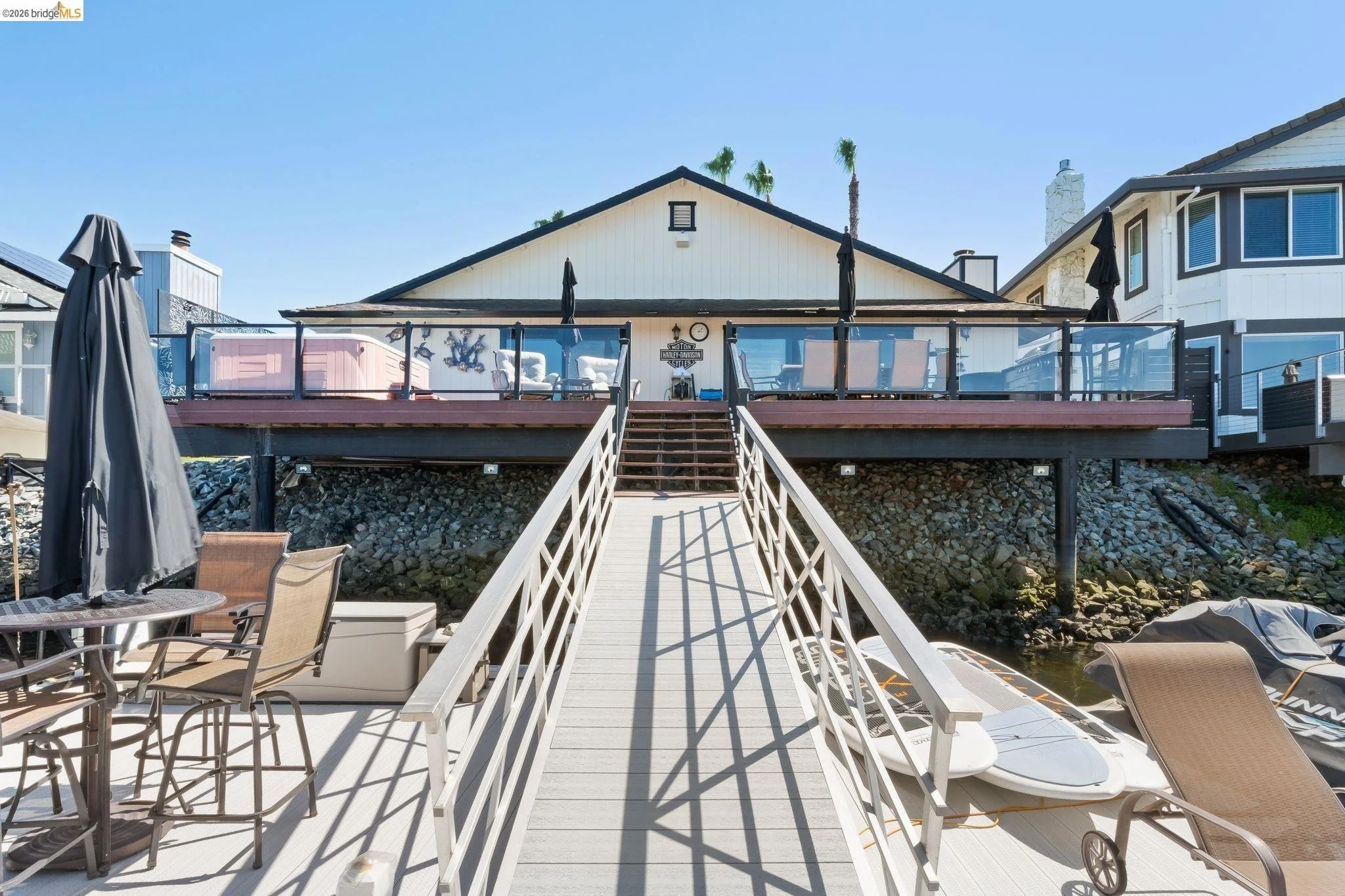View of a house with a large elevated deck overlooking a waterway. The deck has several chairs and umbrellas, a hot tub, and outdoor furniture. A ramp leads up to the deck, with boats docked nearby.