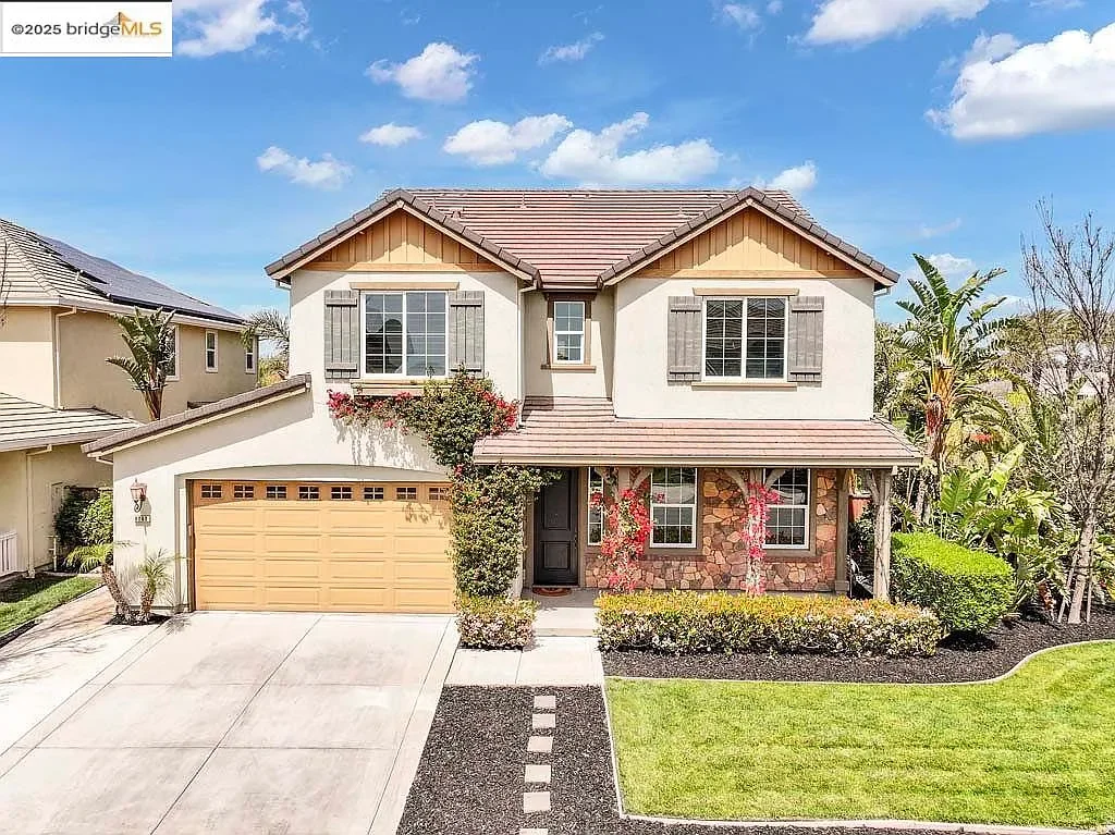 A two-story suburban house with a stone and stucco exterior, a two-car garage, and a well-maintained lawn. The house has gray shutters, a covered porch, and is surrounded by lush greenery and trees under a blue sky with some clouds.