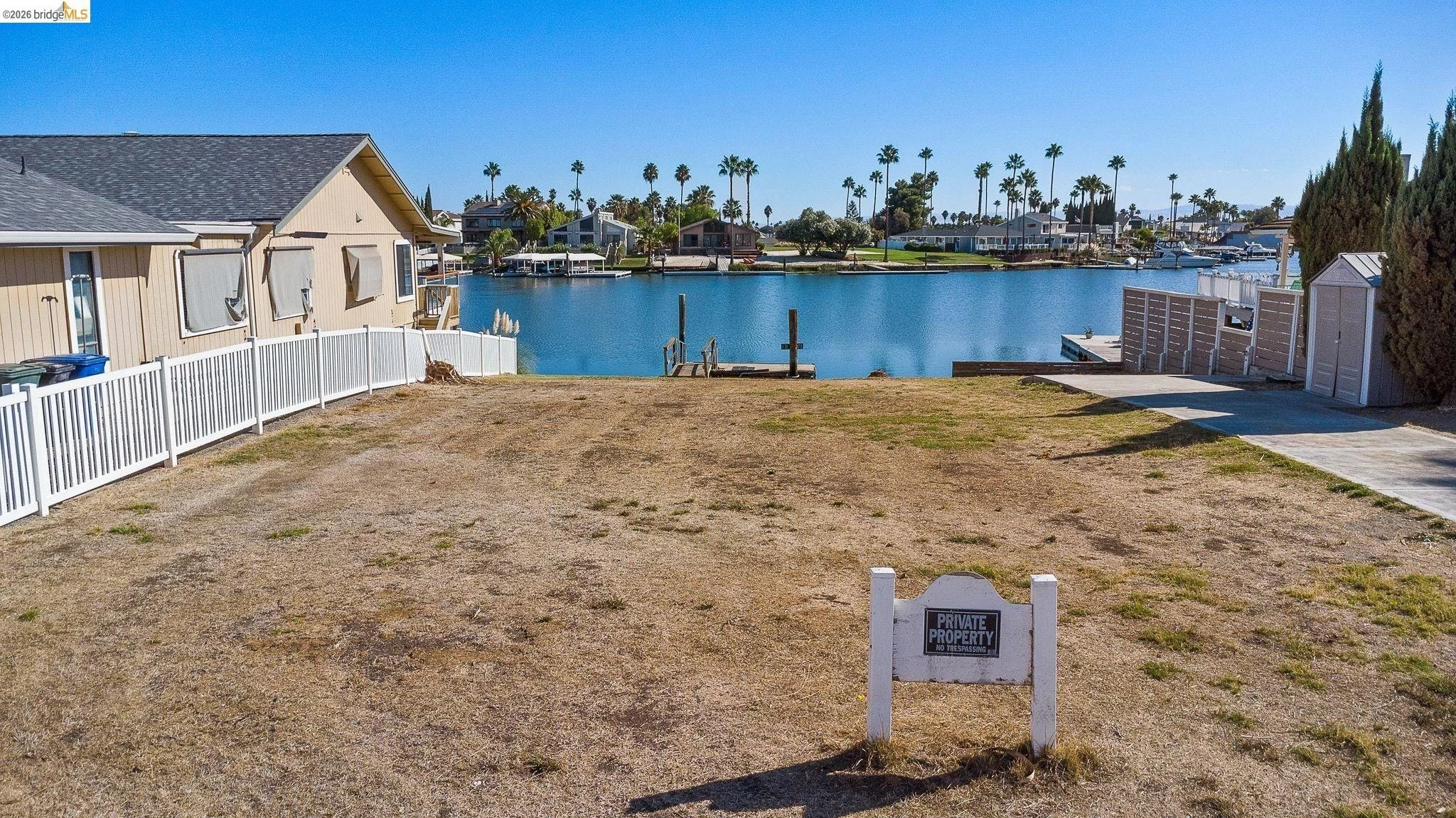 Empty yard with dirt and sparse grass, fenced by a white railing, overlooking a calm waterway with houses and palm trees in the background, and a "Private Property No Trespassing" sign in the foreground.