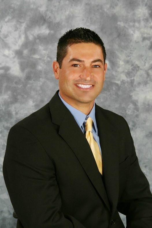 A man in a black suit with a light blue shirt and a yellow tie, smiling, against a gray cloudy background.