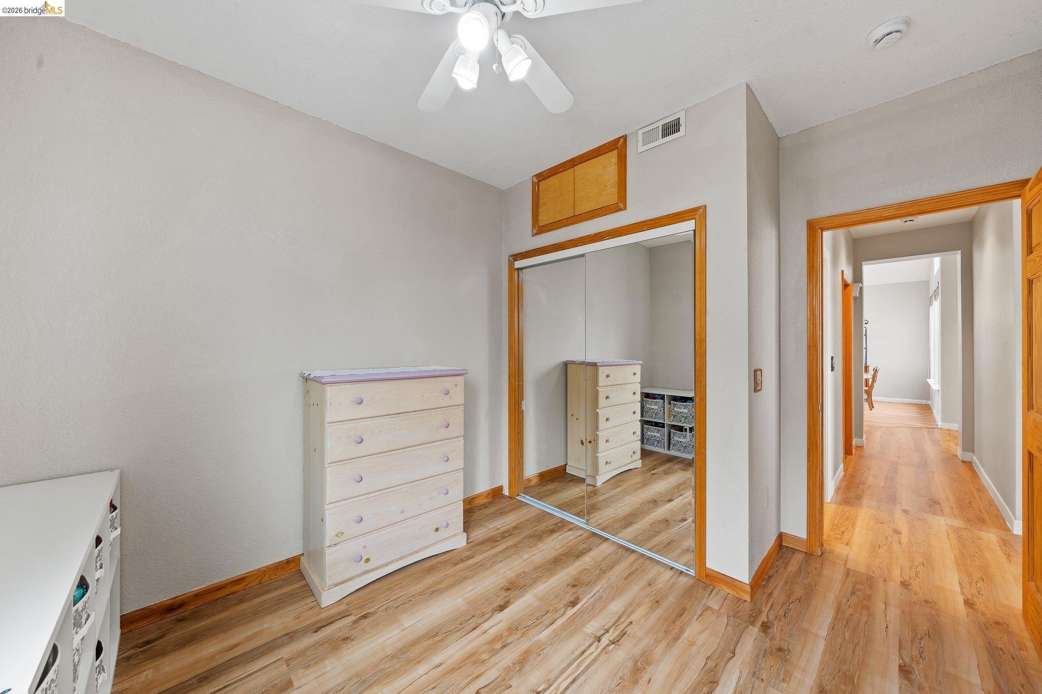 Empty bedroom with hardwood floors, mirrored closet doors, white dressers, and a ceiling fan with lights. A hallway is visible through an open door.