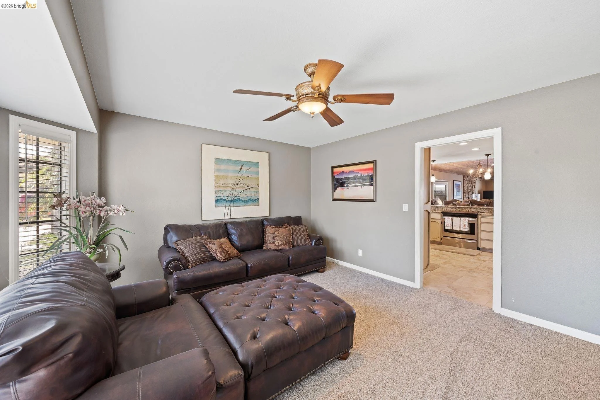 Living room with gray walls, two leather sofas, framed artwork, a ceiling fan, and a window with blinds. The room opens to a kitchen.