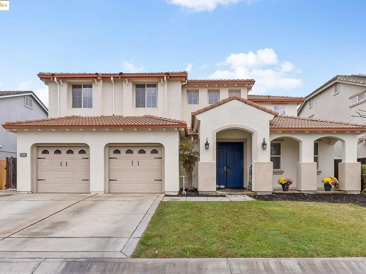 A two-story house with a beige stucco exterior, red roof tiles, and a dark blue front door. The house features two garage doors, and the front yard has a small patch of grass with two flower pots and a small tree.