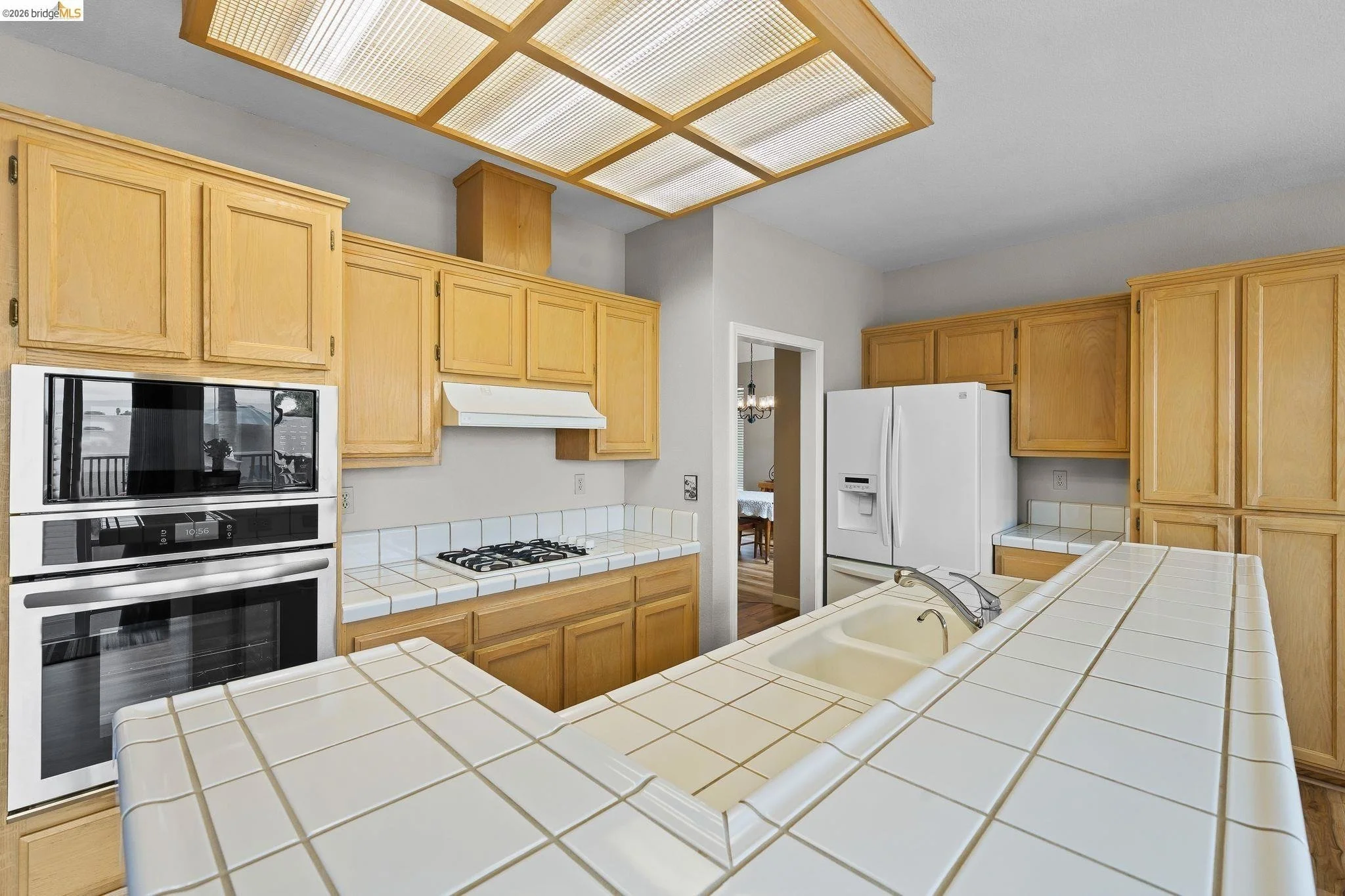 Kitchen with wooden cabinets, white tile countertops, a built-in oven, a stovetop, a white refrigerator, and a central sink with an island counter.