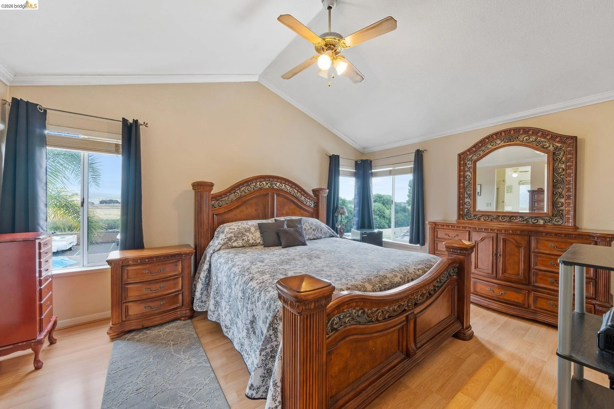 Bedroom with wooden bed frame, matching dresser with mirror, nightstand, and blue curtains on windows, ceiling fan with light, beige walls, and wood flooring.