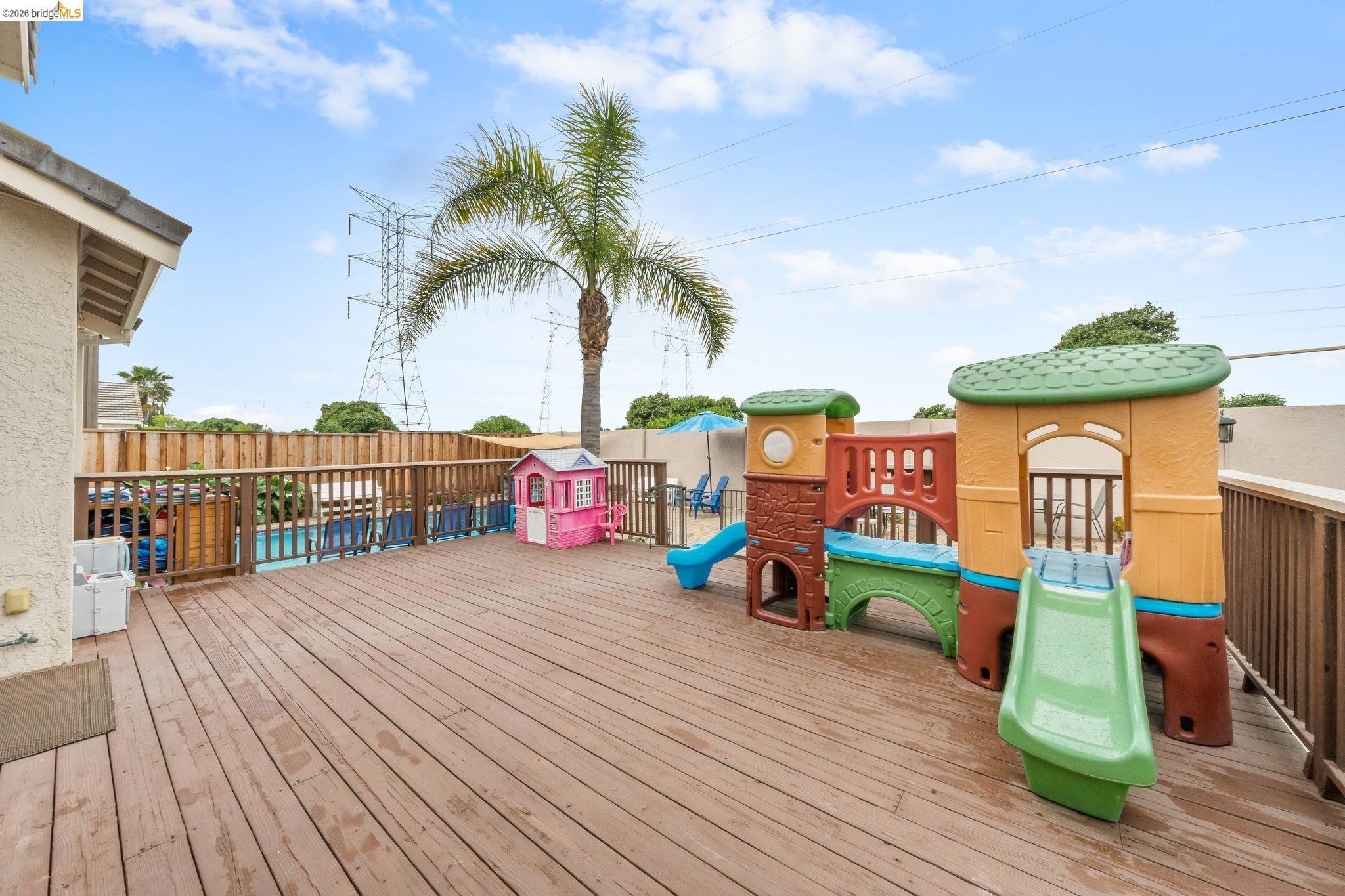 Wooden decked backyard with colorful plastic play structures, including slides and a playhouse, a palm tree, and clear sky with some clouds.