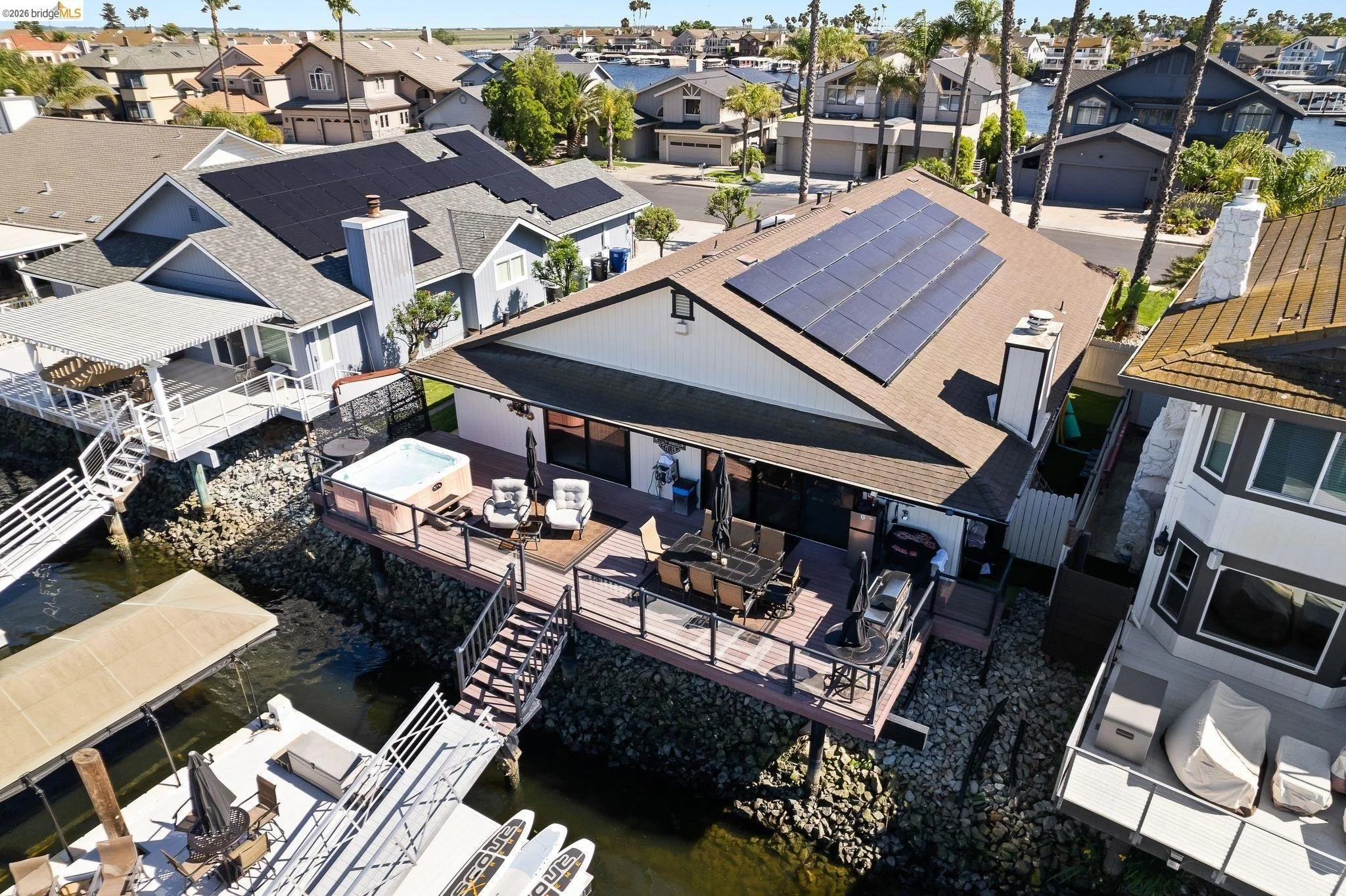 Aerial view of a waterfront house with a deck, outdoor furniture, hot tub, and solar panels on the roof, situated in a neighborhood with similar houses and palm trees.