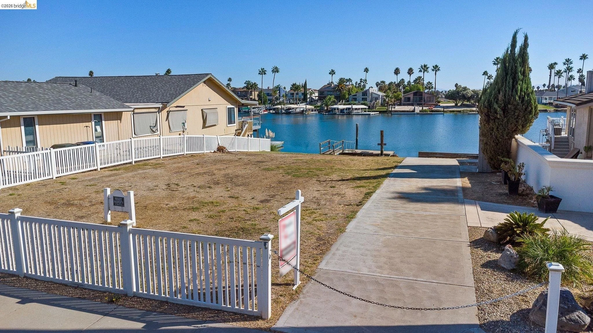 View of a fenced yard with a sidewalk leading to a lake, houses, and palm trees in the background.