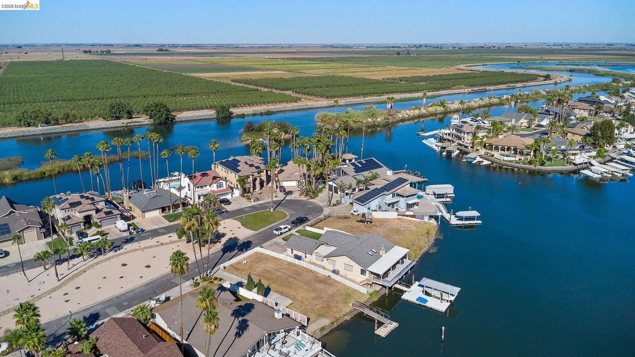 Aerial view of waterfront houses with boats, surrounded by a large river, with agricultural fields and farmland in the background.