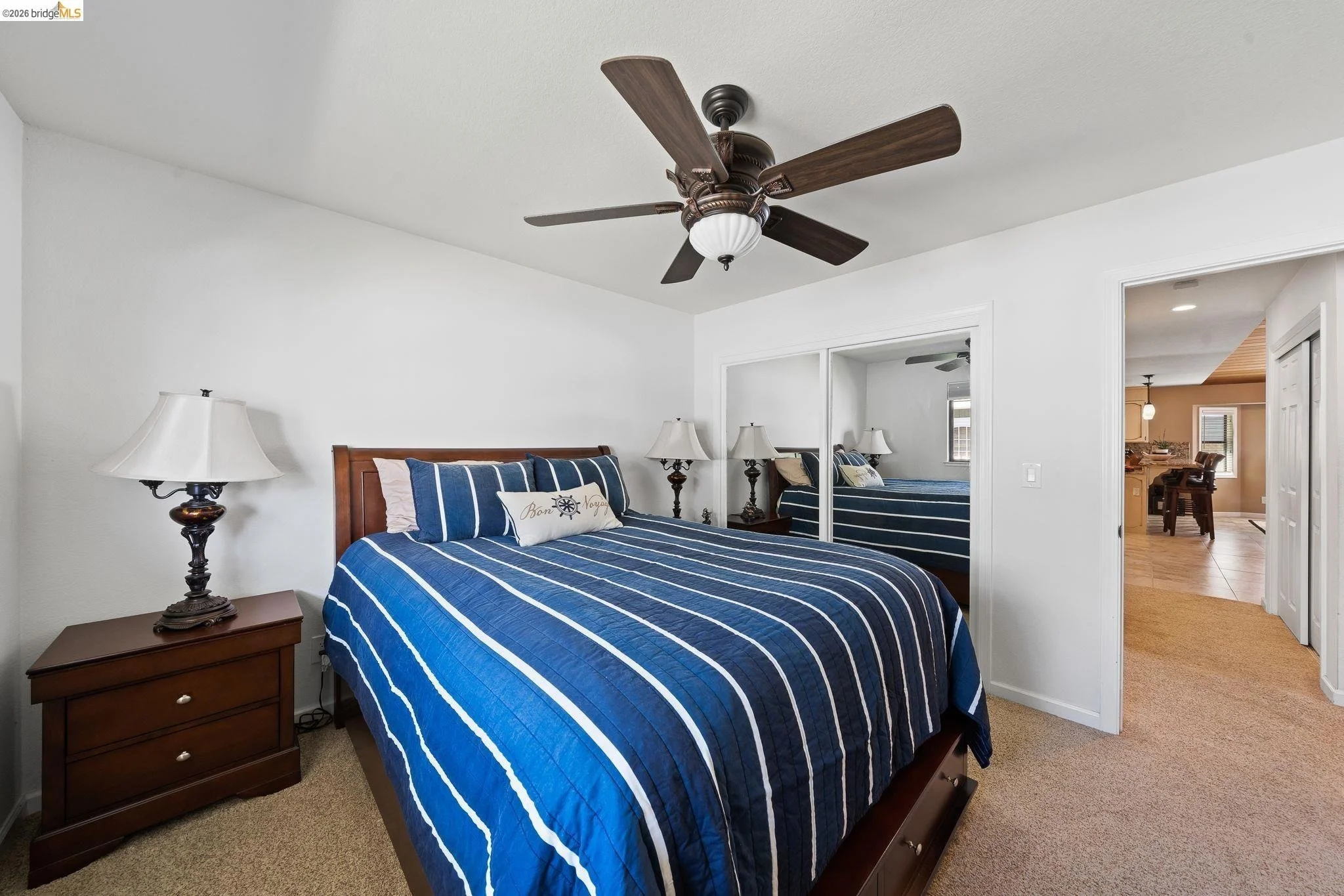Bedroom with a wooden bed frame, blue striped bedding, two bedside tables with lamps, ceiling fan, mirrored closet door, and view into dining area.