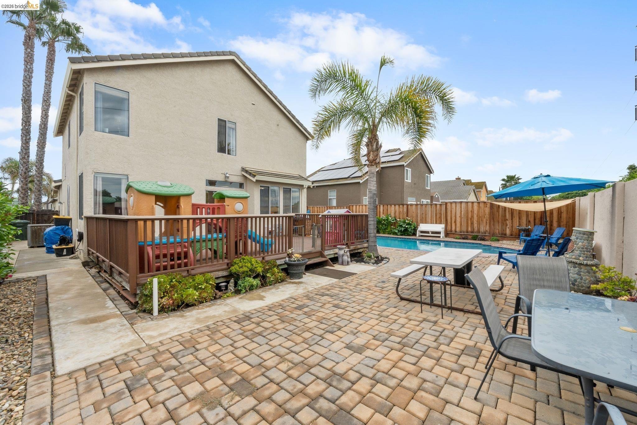 Backyard with swimming pool, patio area with tables and chairs, children's playset, palm trees, and fenced yard.