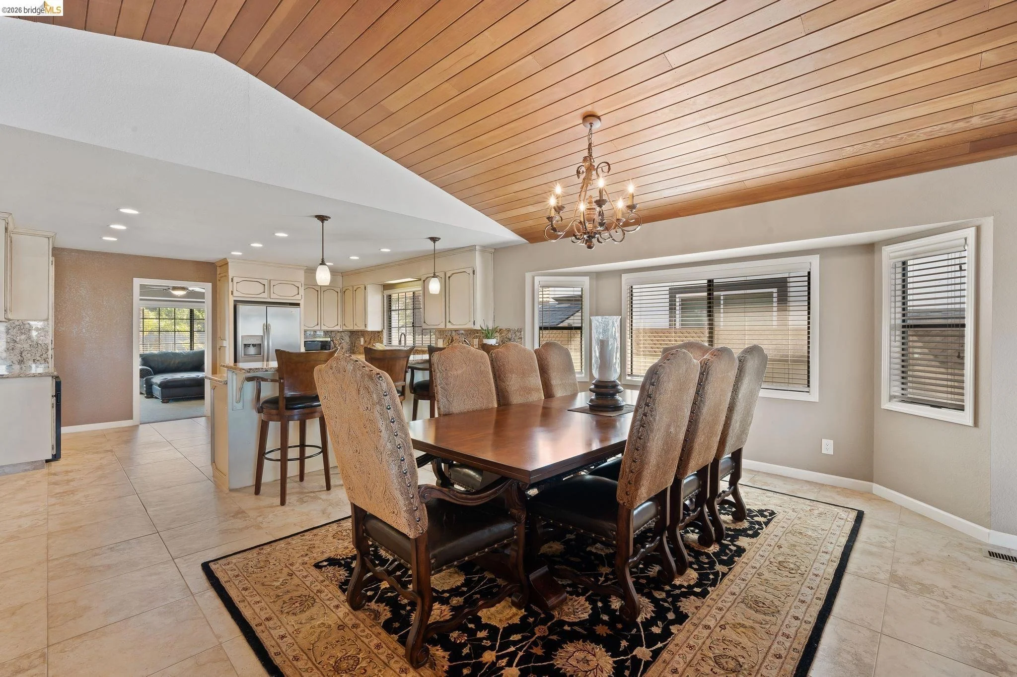 Dining room with a wooden table, eight upholstered chairs, a chandelier, windows with blinds, and a view into the kitchen and living room.