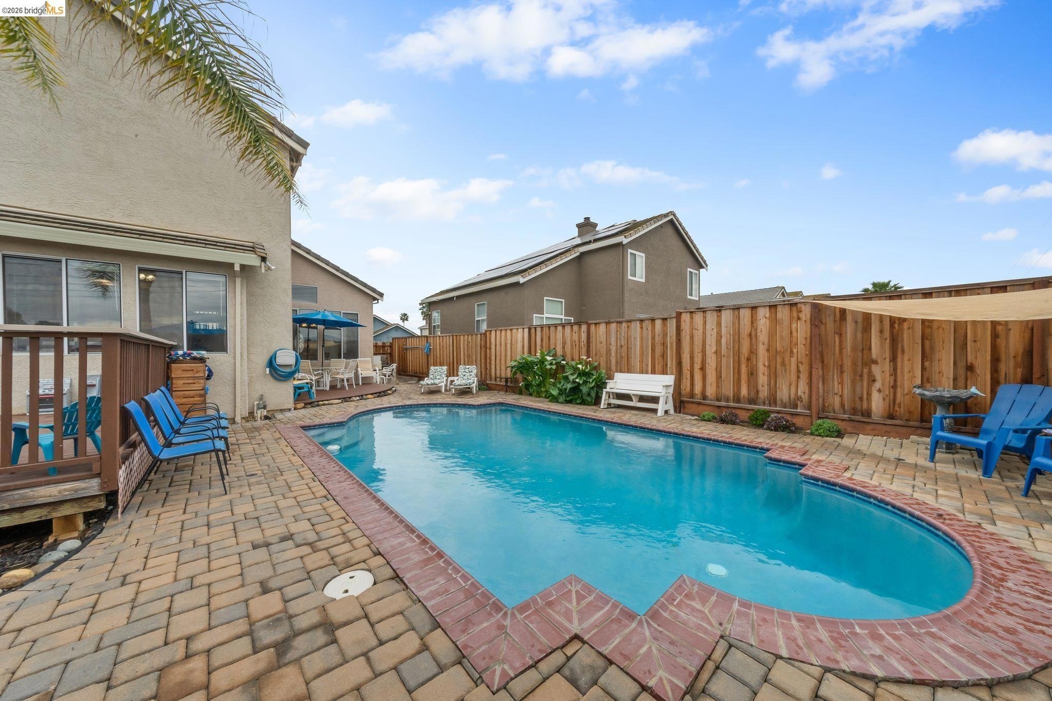 Residential backyard with a swimming pool, patio furniture, potted plants, and a wooden fence under a partly cloudy sky.