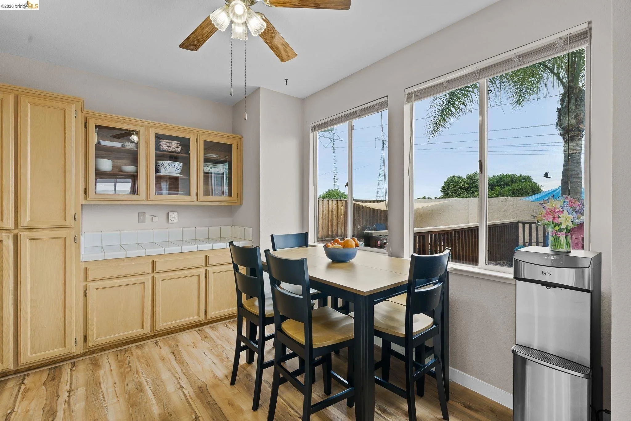 A kitchen dining area with a wooden table and black chairs, large windows overlooking a backyard with trees, a palm tree, and a fence, a water dispenser, and ceiling fan with lights.