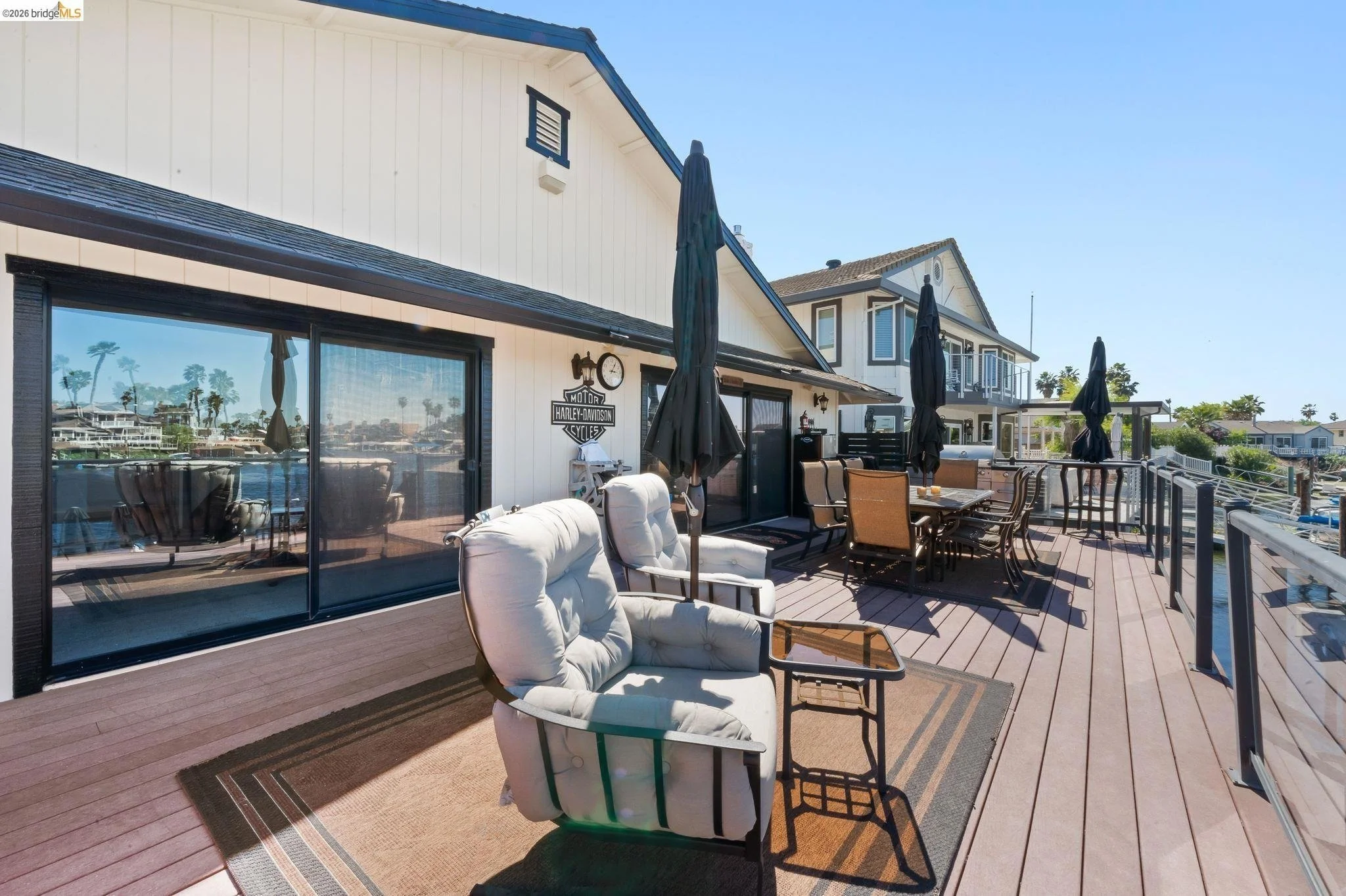 Large wooden deck on a waterfront house with outdoor furniture including cushioned chairs, a dining table with multiple chairs, large black umbrellas, and a glass railing. Reflected palm trees and neighboring houses are visible in the sliding glass d