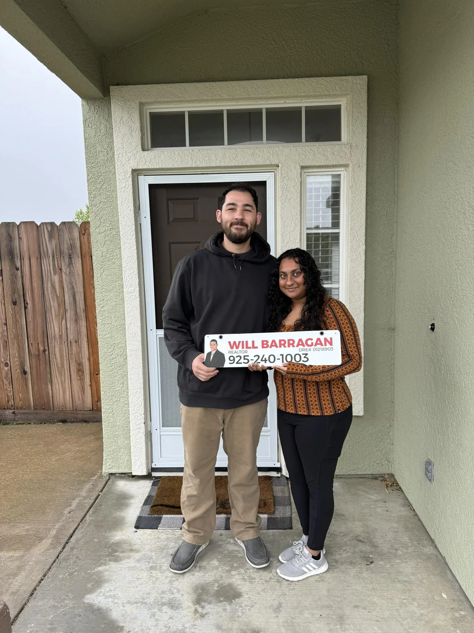 A man and woman standing in front of a house door, holding a sign with real estate information, including a photo of the man, the name 'Will Barragan,' and a phone number.