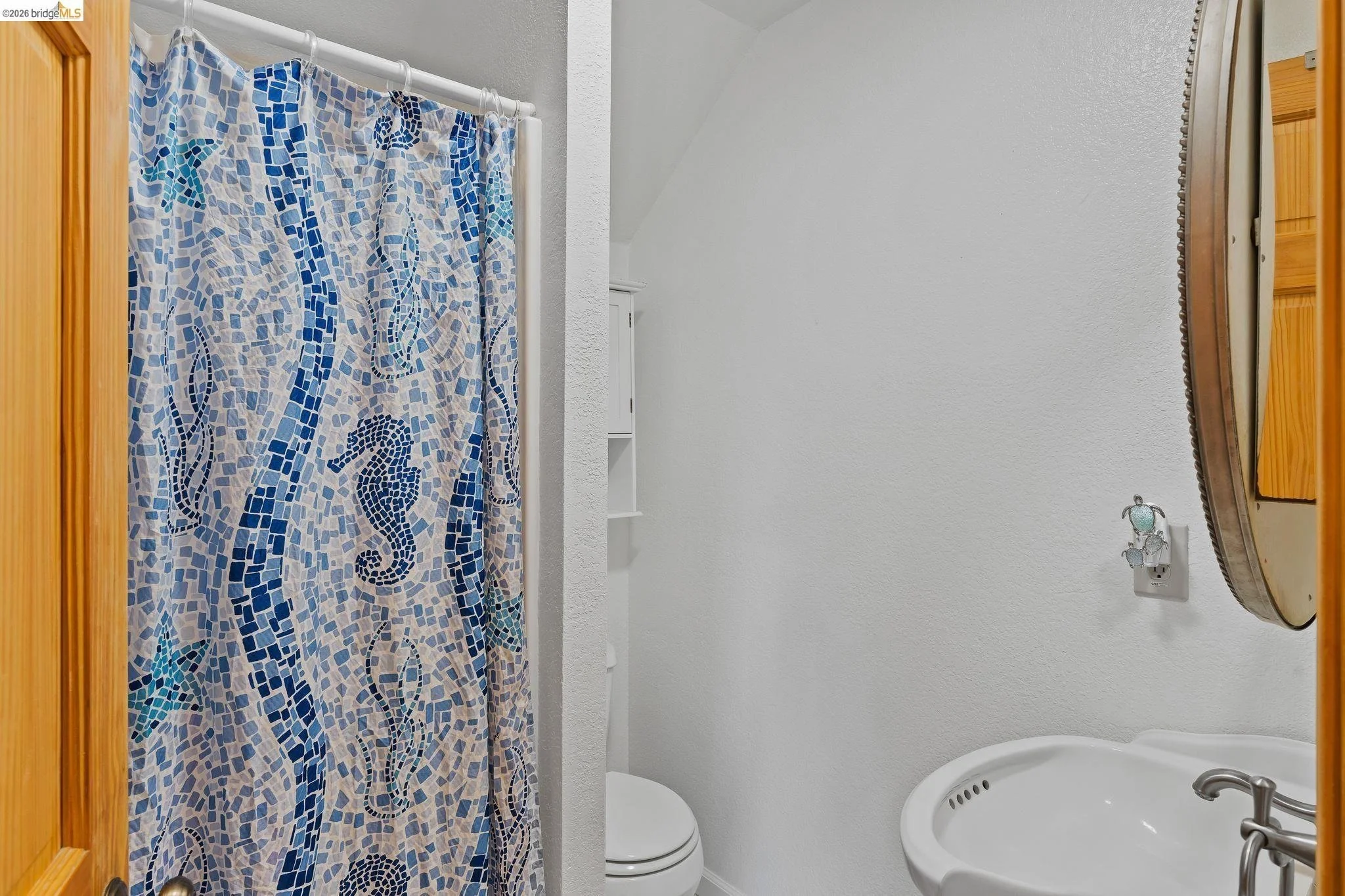 A small bathroom with a shower curtain depicting a mosaic seahorse design in blue tones, a white toilet, a curved mirror above a white sink, and a light-colored wooden door frame.