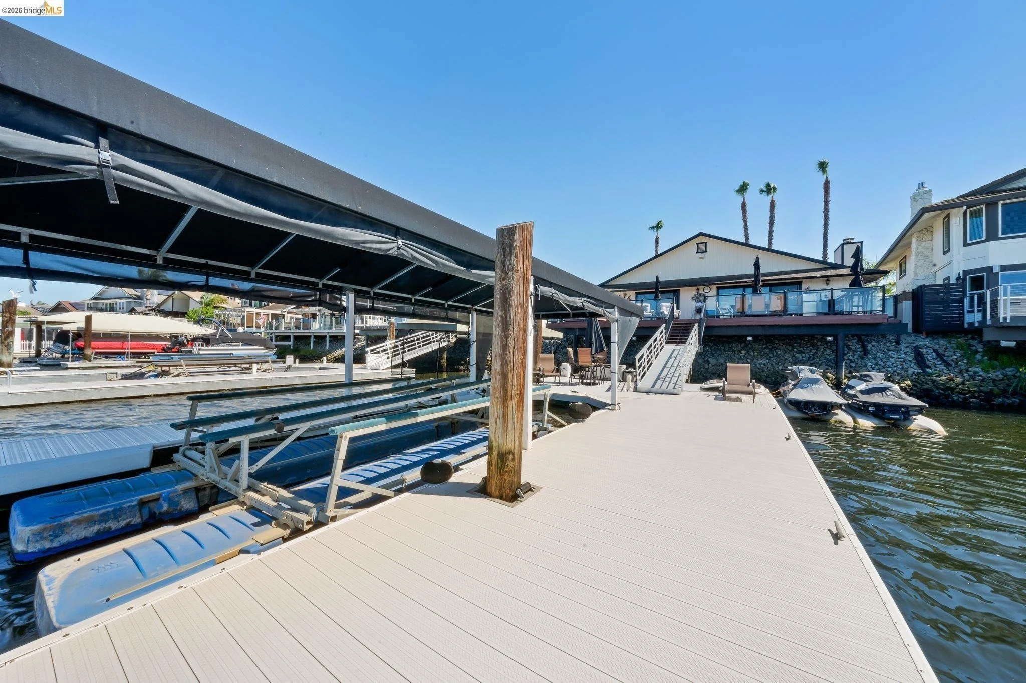 Boat dock with covered boat slips, watercraft, and residential houses with palm trees in the background under a clear blue sky.