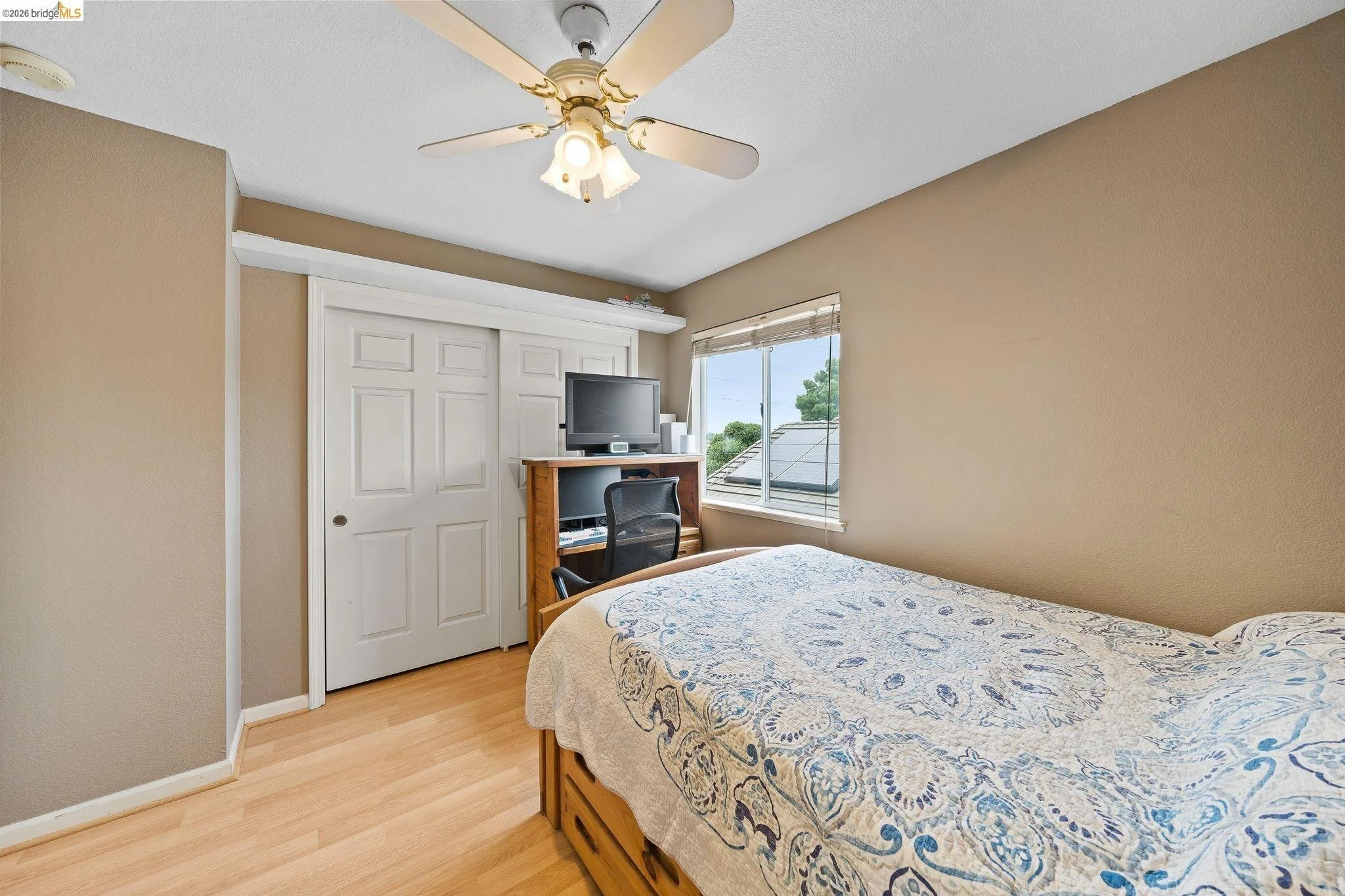 A bedroom with a bed, a closet with sliding doors, a desk with a computer monitor, chair, and window showing rooftops and trees outside.