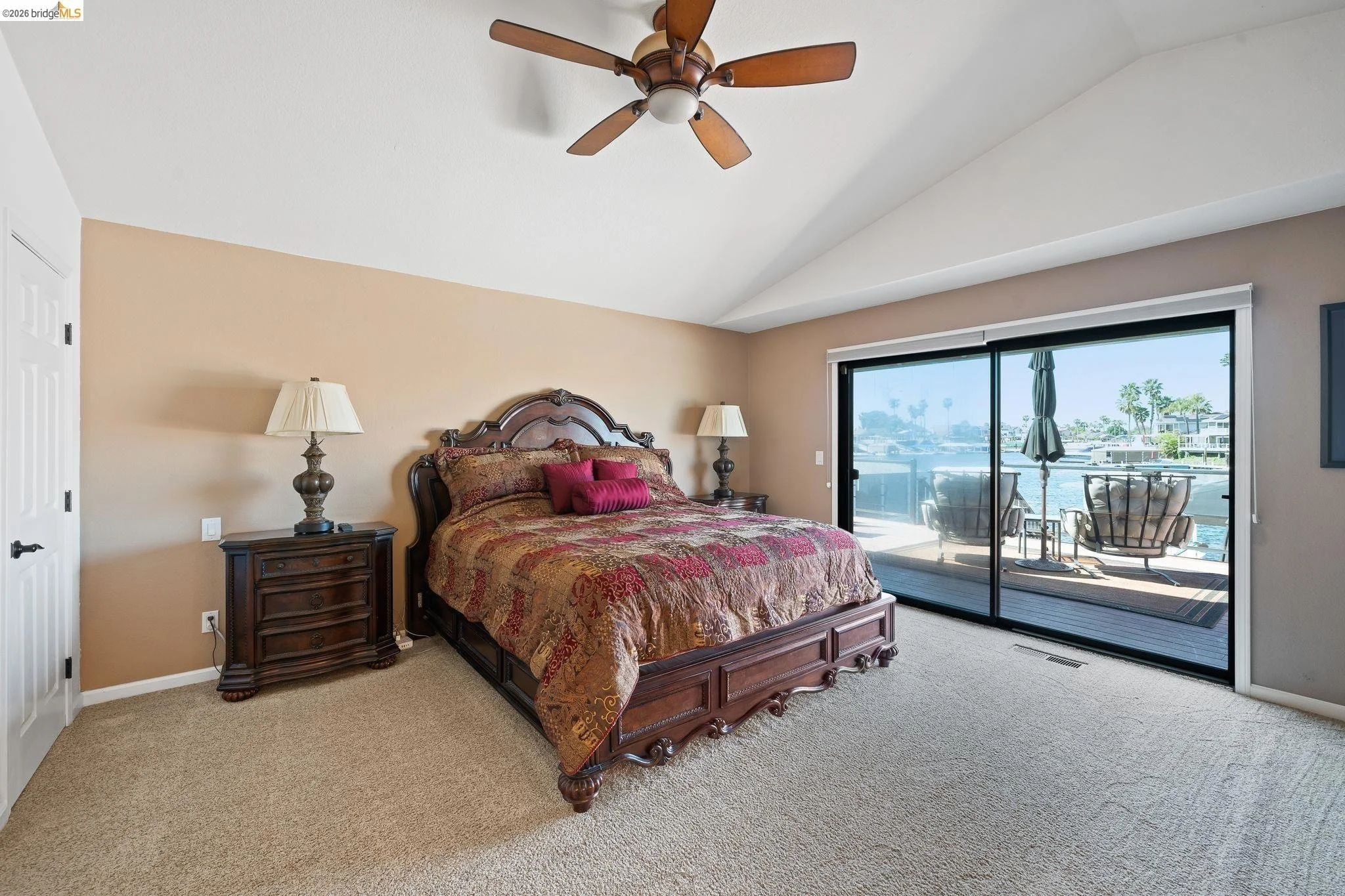 Bedroom with a large wooden bed, beige walls, and a sliding glass door leading to a balcony overlooking a waterway with boats, patio furniture, and palm trees.