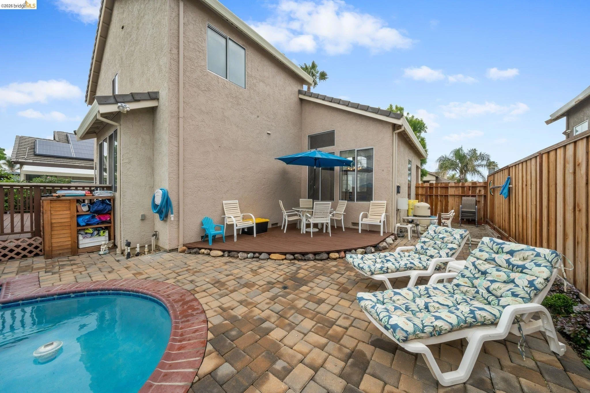 Backyard patio with pool, Sun loungers with floral cushions, patio table with chairs and blue umbrella, wooden deck, and fenced yard, house with beige stucco walls and large windows, cloudy sky