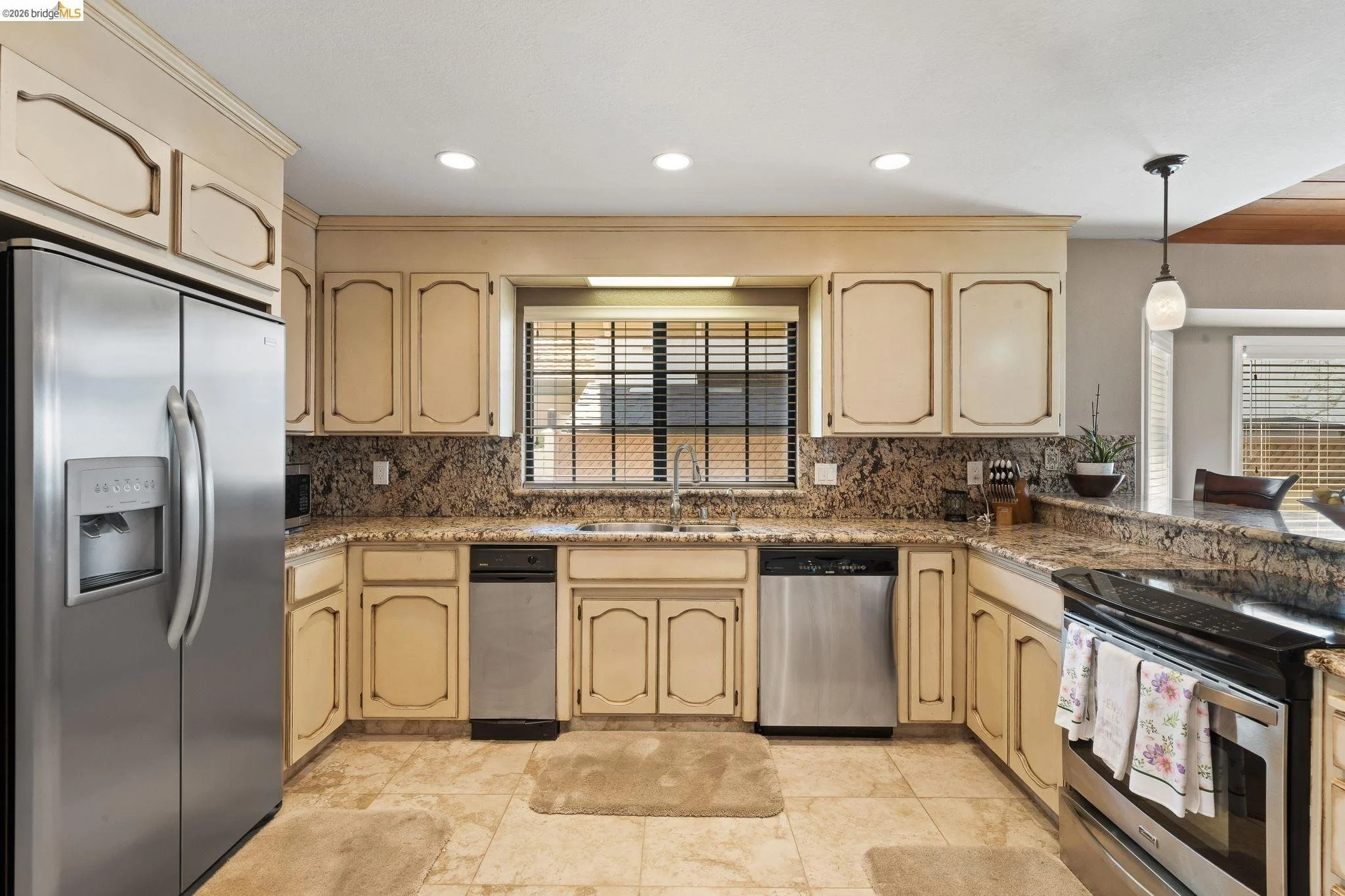Kitchen with beige cabinets, granite countertops, stainless steel refrigerator, dishwasher, and oven. There is a window above the sink with blinds, and a small potted plant on the counter.