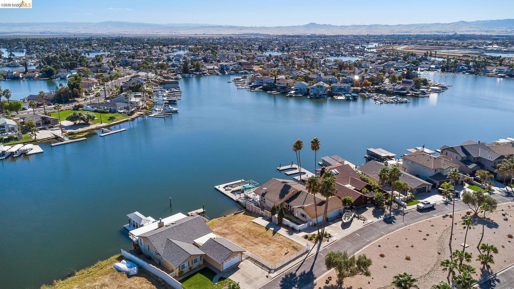 Aerial view of a coastal residential neighborhood with waterfront houses, boats, and palm trees, with a large body of water and distant hills in the background.