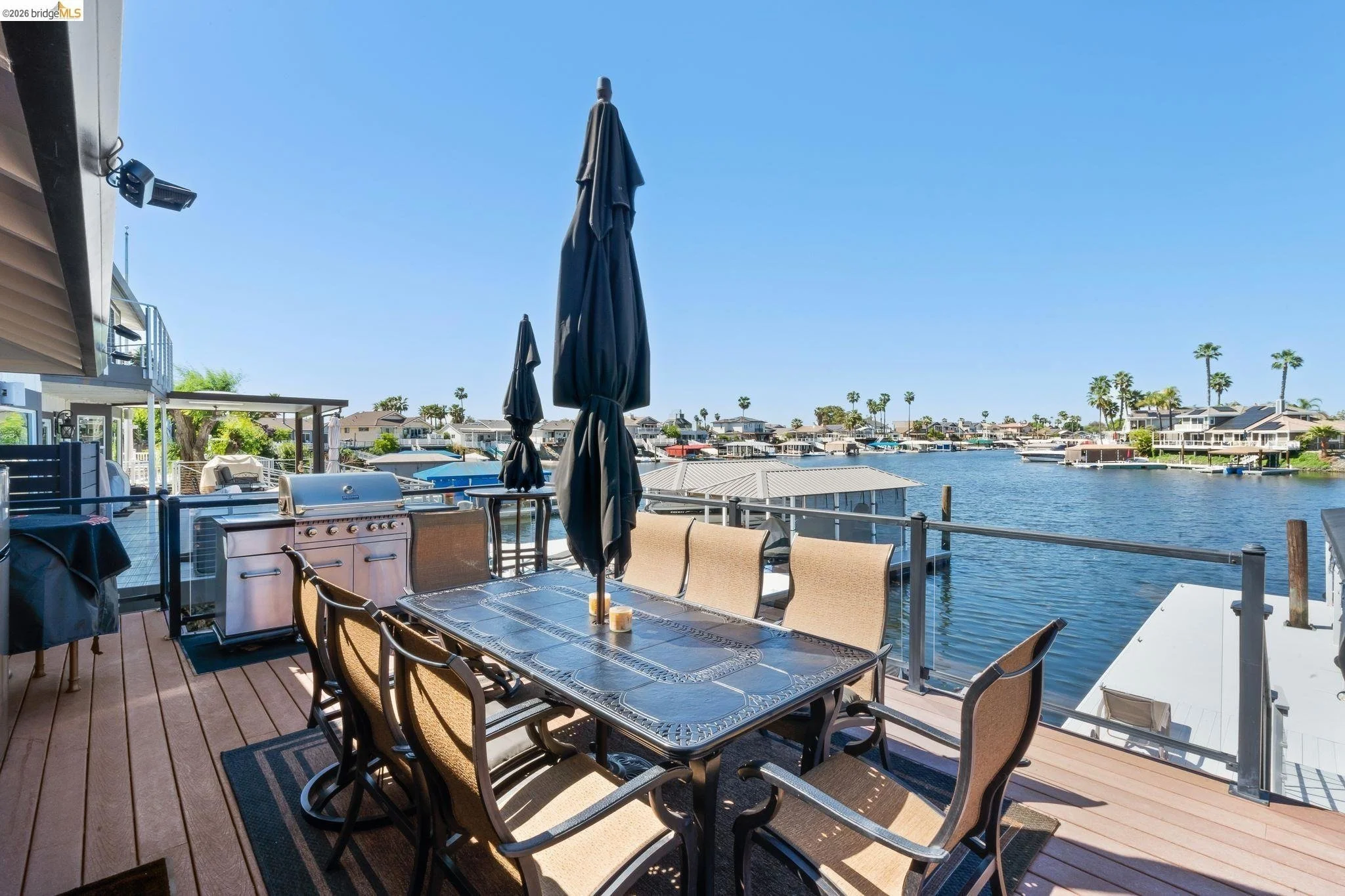 A waterfront outdoor patio with a dining table, six chairs, a barbecue grill, and a large black umbrella. The patio overlooks a calm waterway with boats, houses, palm trees, and a clear blue sky in the background.
