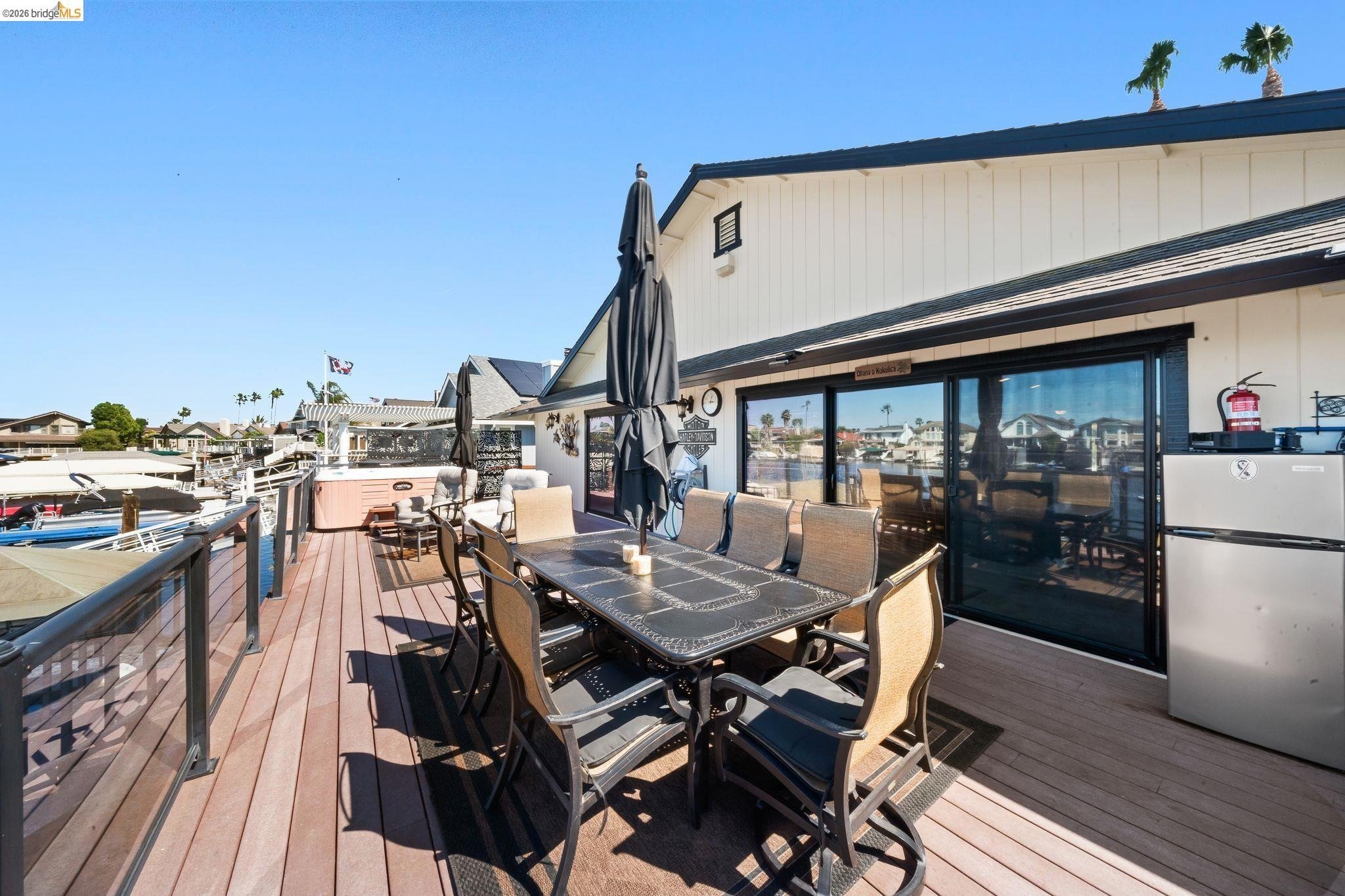 Empty outdoor deck with dining table, chairs, and large umbrellas overlooking a marina with boats and water in the background.
