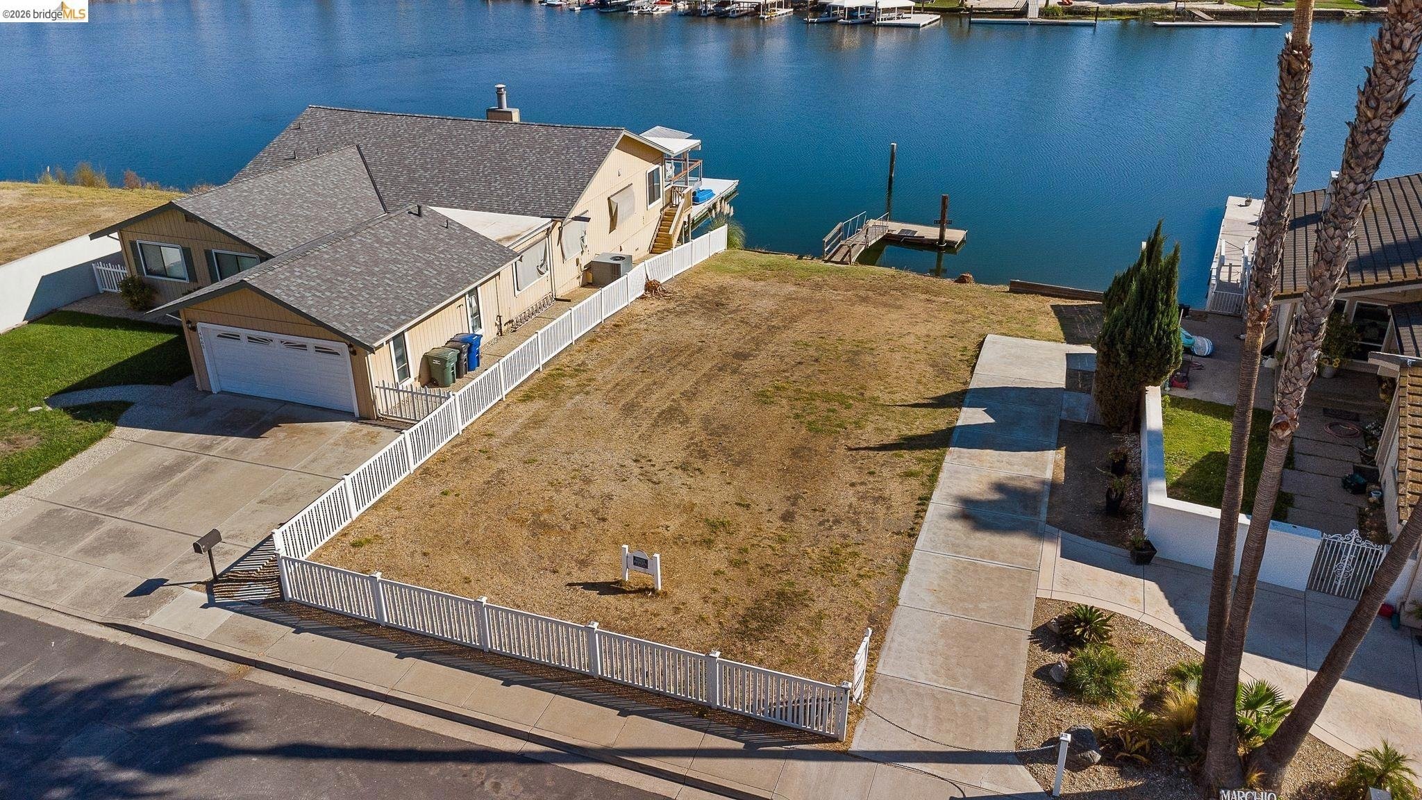 Aerial view of a waterfront property featuring a house with a garage, a fenced yard with dry grass, a concrete driveway, a sidewalk, and a marina with boats on a river or lake in the background.