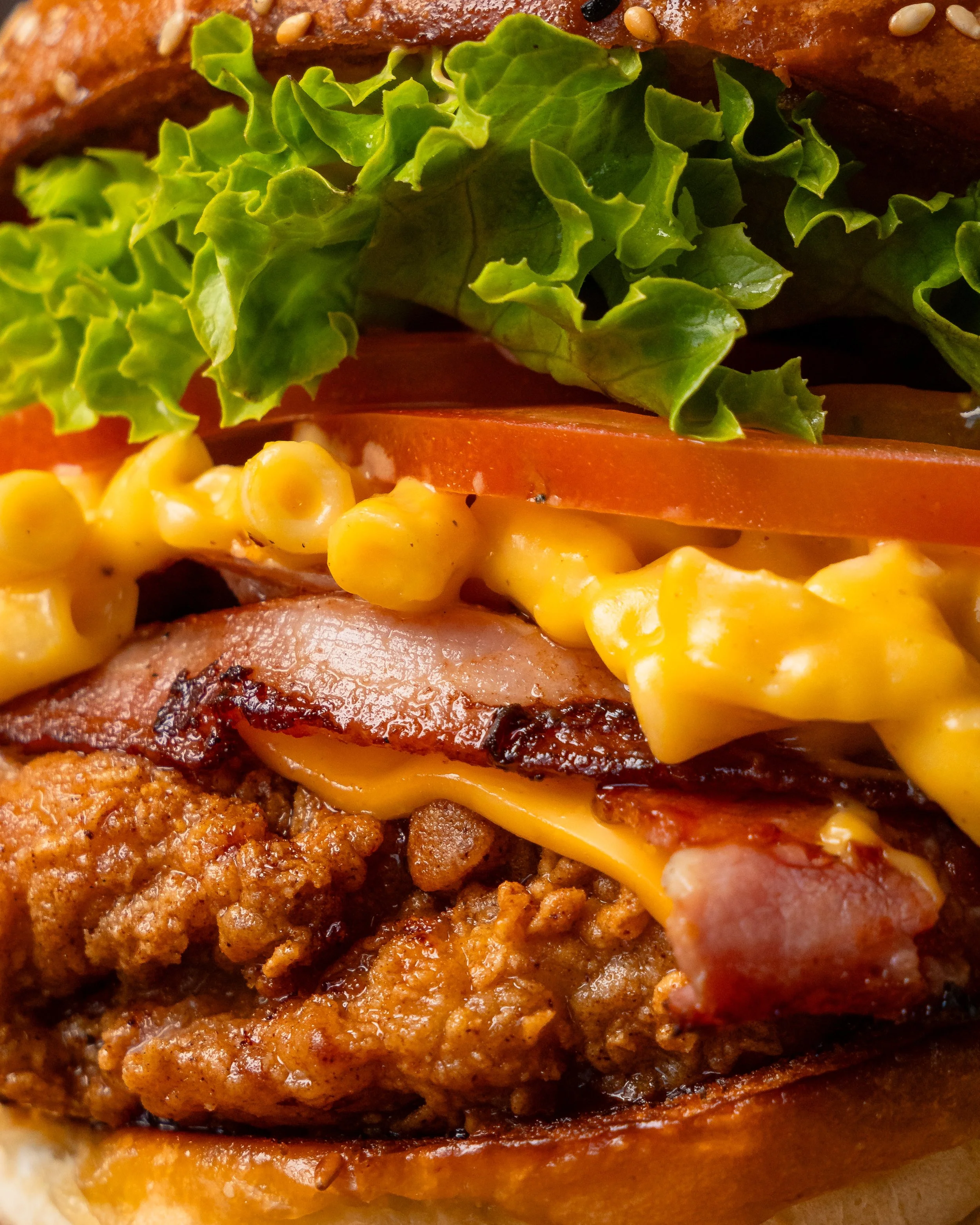 Close-up of a burger with fried chicken patty, cheddar cheese, bacon, tomato slices, lettuce, and macaroni and cheese on a bun.