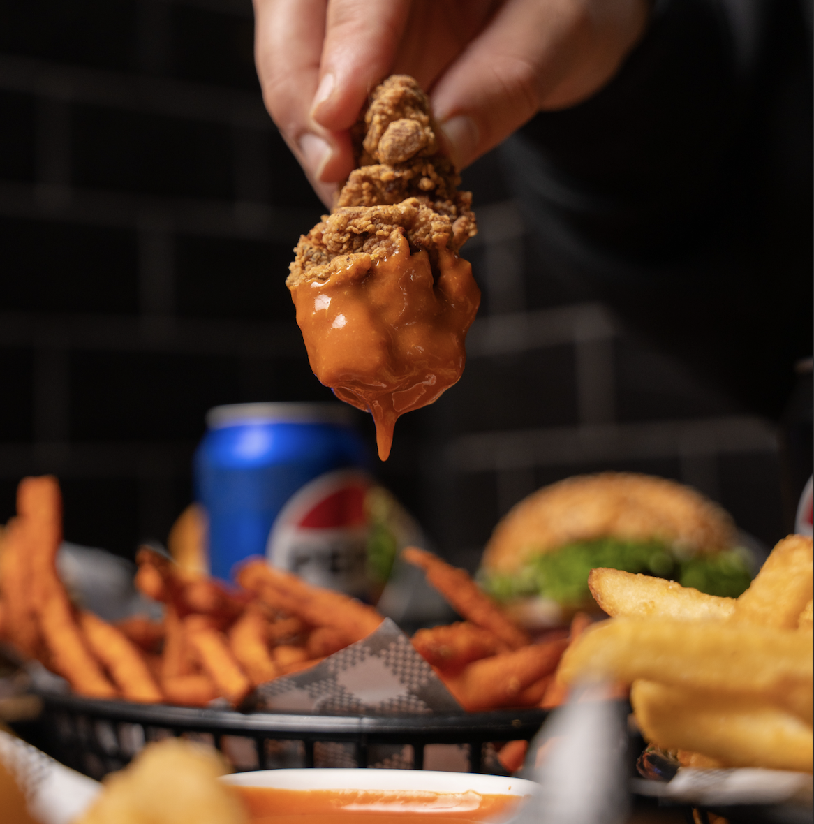 A hand holding a piece of fried chicken dipped in orange sauce over a basket of sweet potato fries, with a burger and a can of Pepsi in the background.