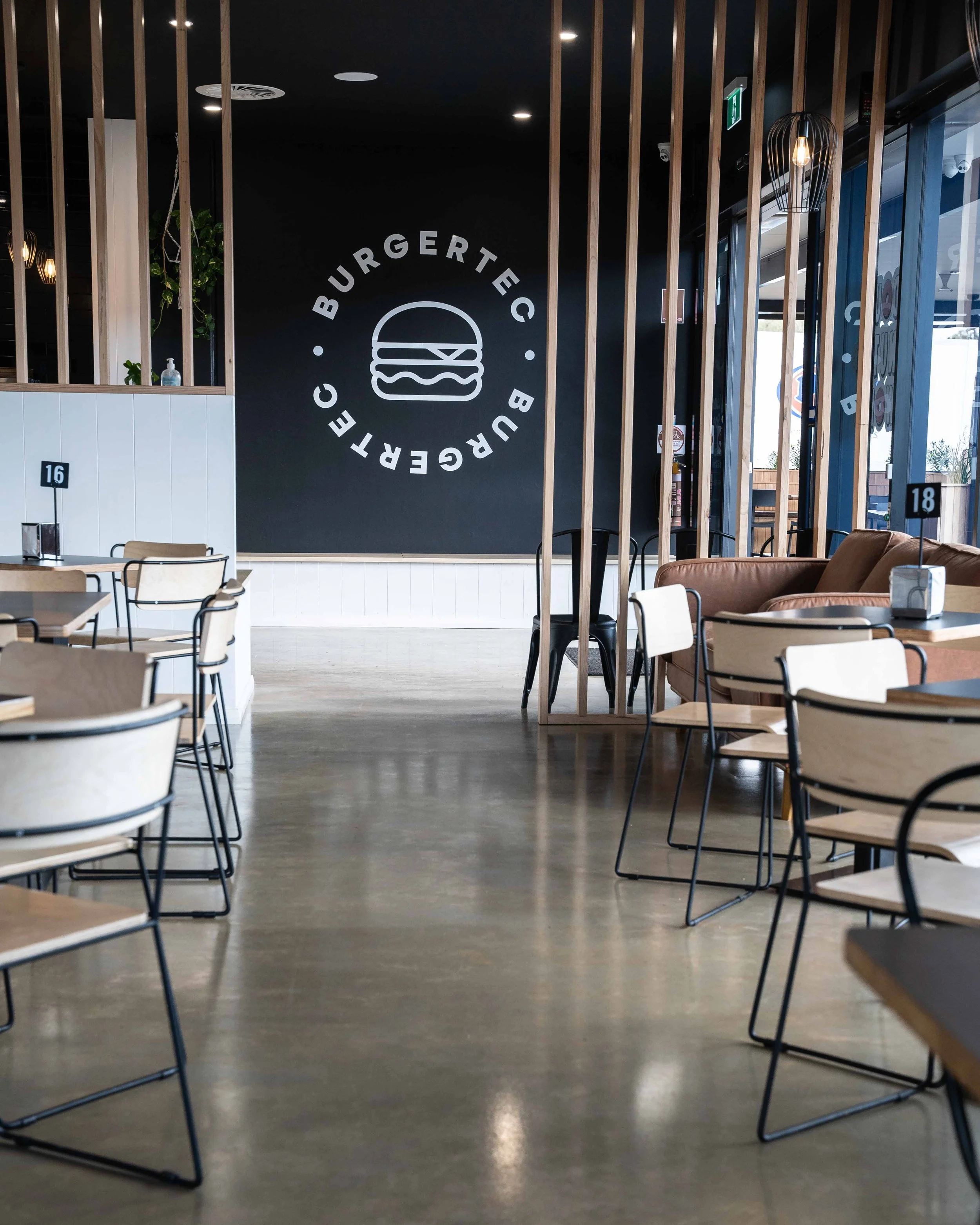 Interior of a modern burger restaurant with wooden tables and chairs, a black wall with cartoon burger logo and the words "BURGERTEC BC", and a leather couch near the window.