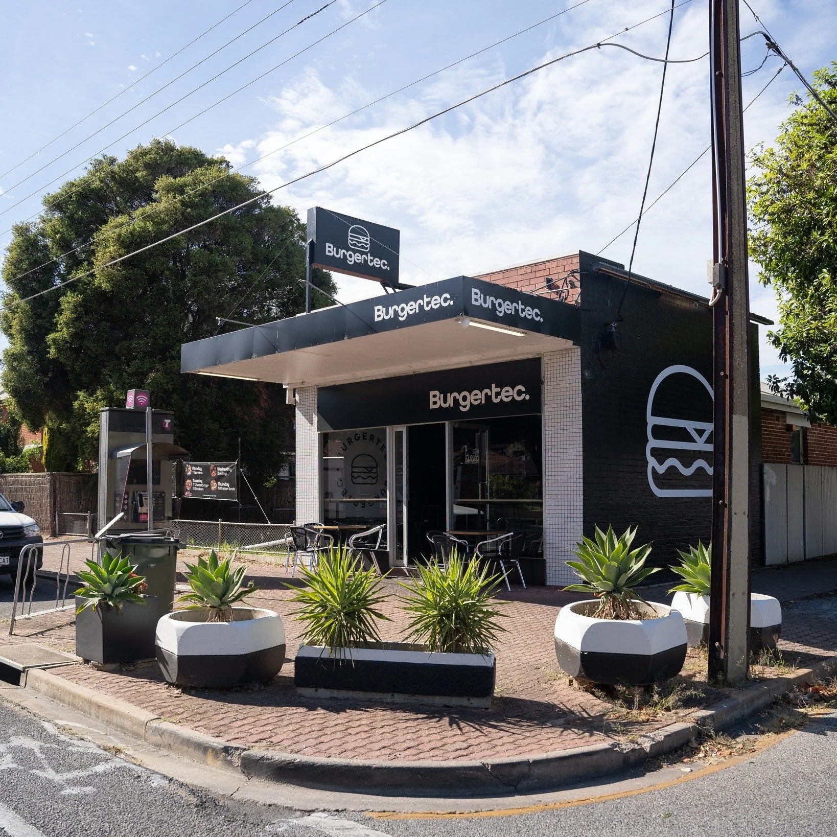 Exterior of a burger restaurant named 'Burgertec,' with a black and white color theme, outdoor seating, potted plants, and a large burger logo on the side of the building.