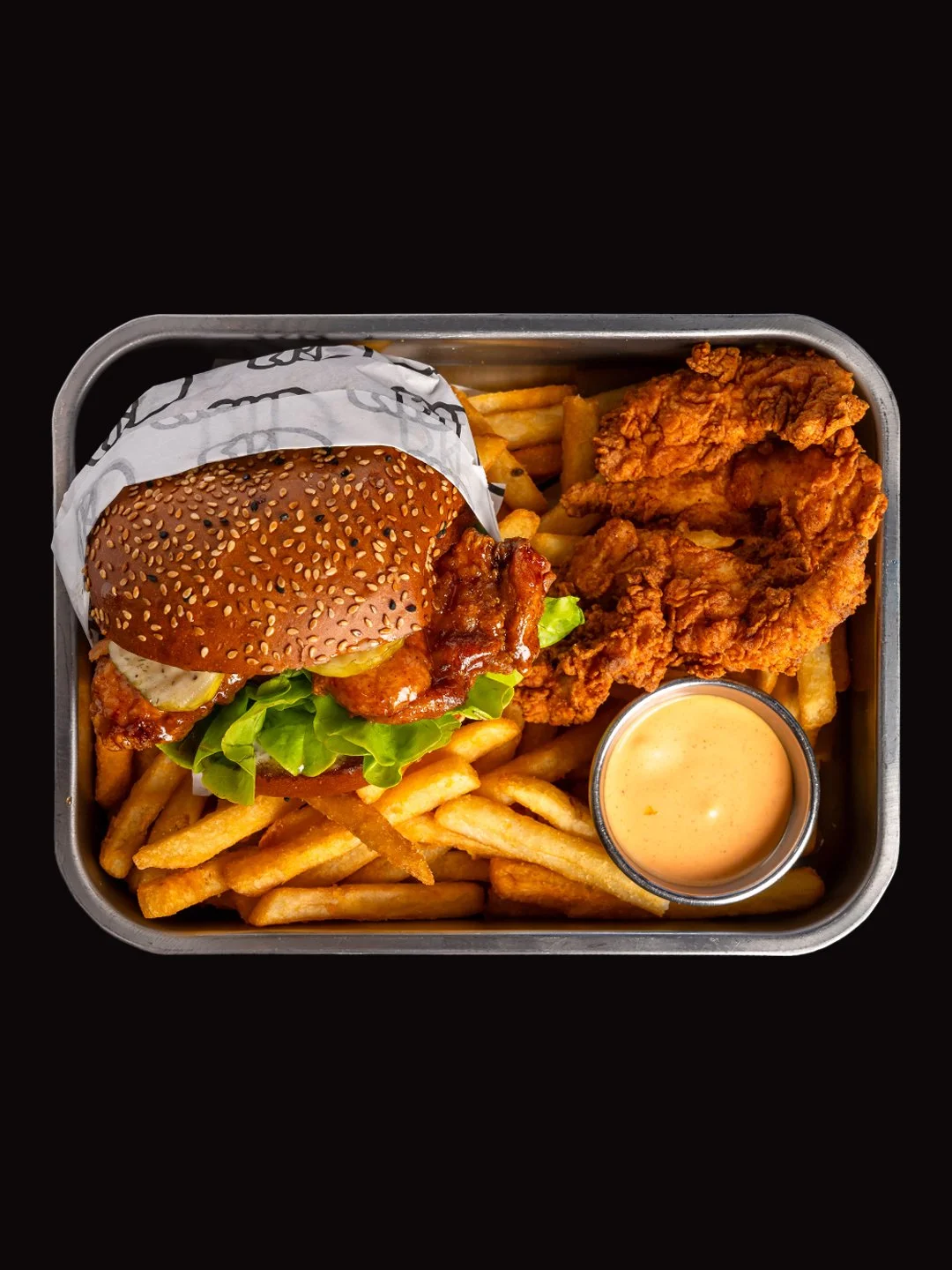 A metal tray of fast food containing a sesame seed burger with lettuce, tomato, pickles, fried chicken, French fries, and a cup of orange dipping sauce.
