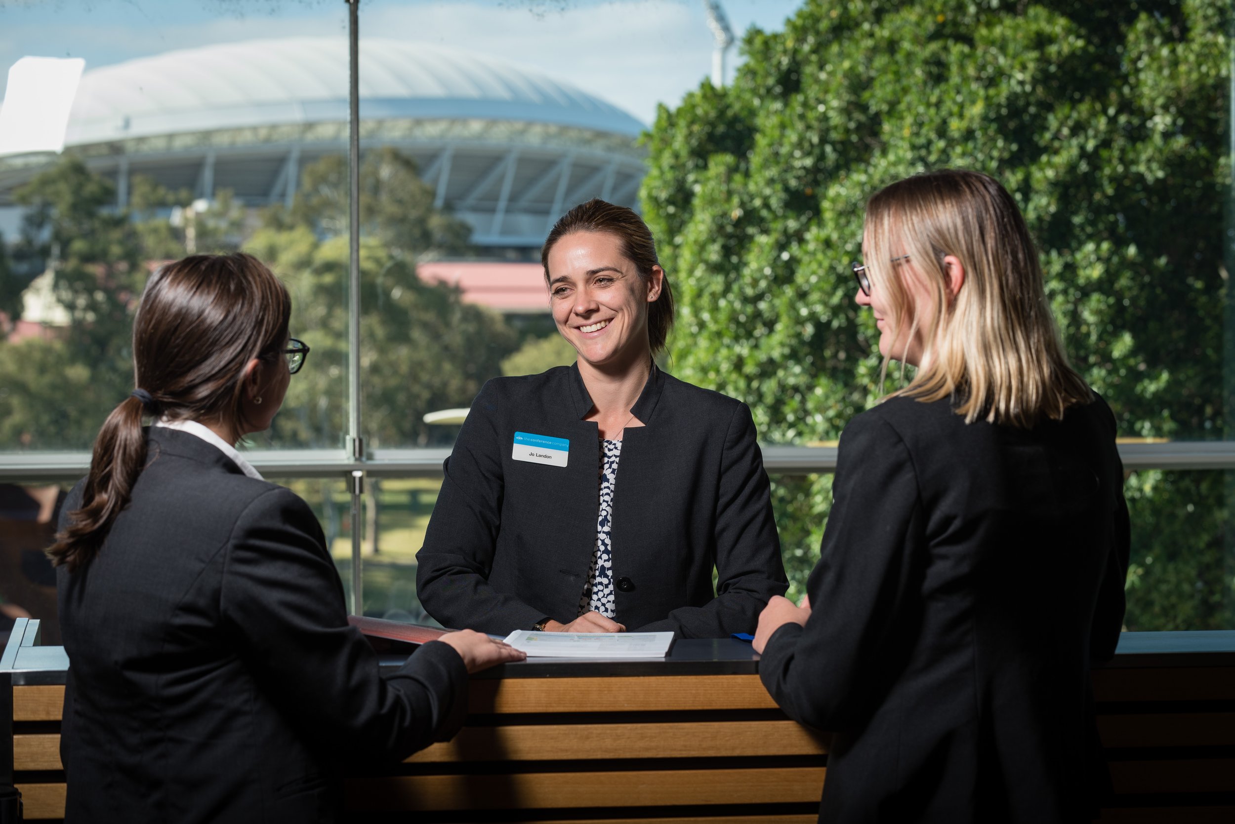 Three women in business attire having a conversation during a meeting, seen through a glass window with green trees and a stadium in the background.