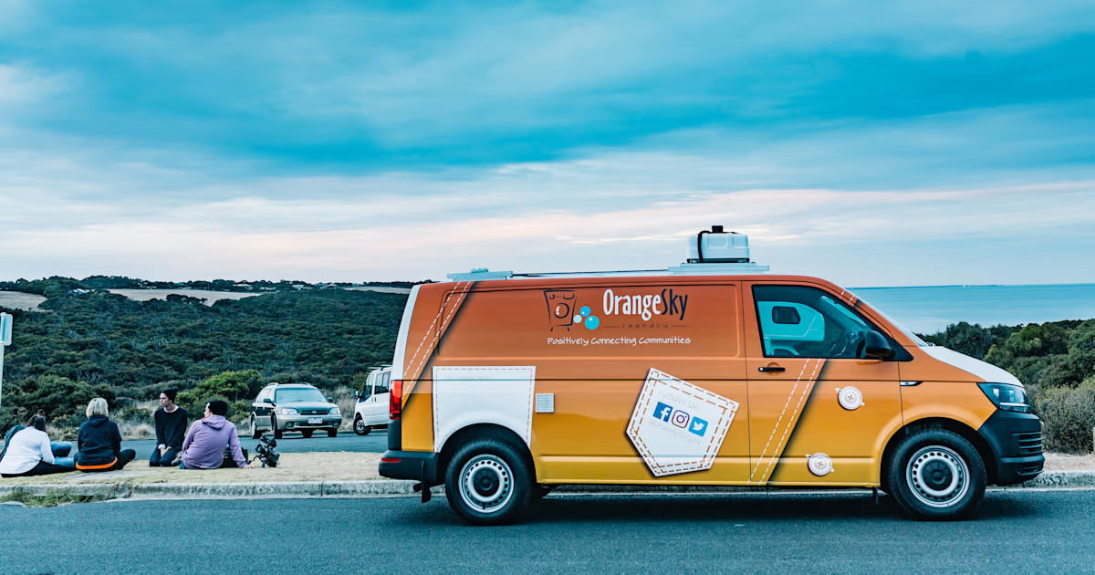 Orange Sky laundry van parked on side of road with four people sitting on curb and trees in background.