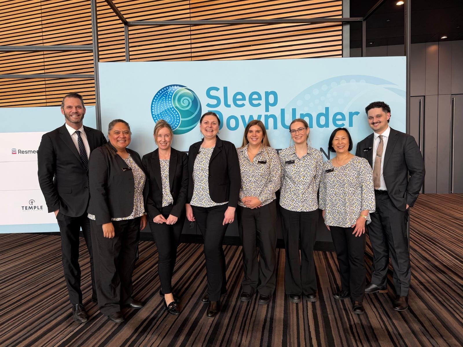 Group of eight professionally dressed people standing in front of a sign that reads 'Sleep Downunder,' in a modern building with wood paneling and a striped carpet.