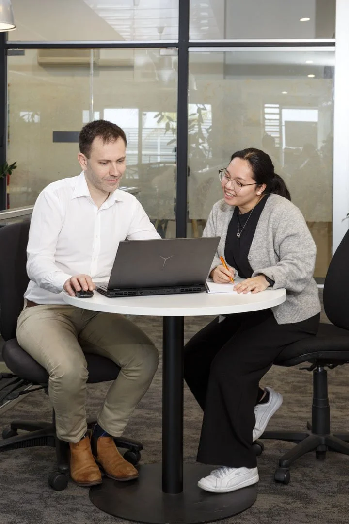 Two people sitting at a white round table in an office, looking at a laptop, smiling, with a pen in hand.