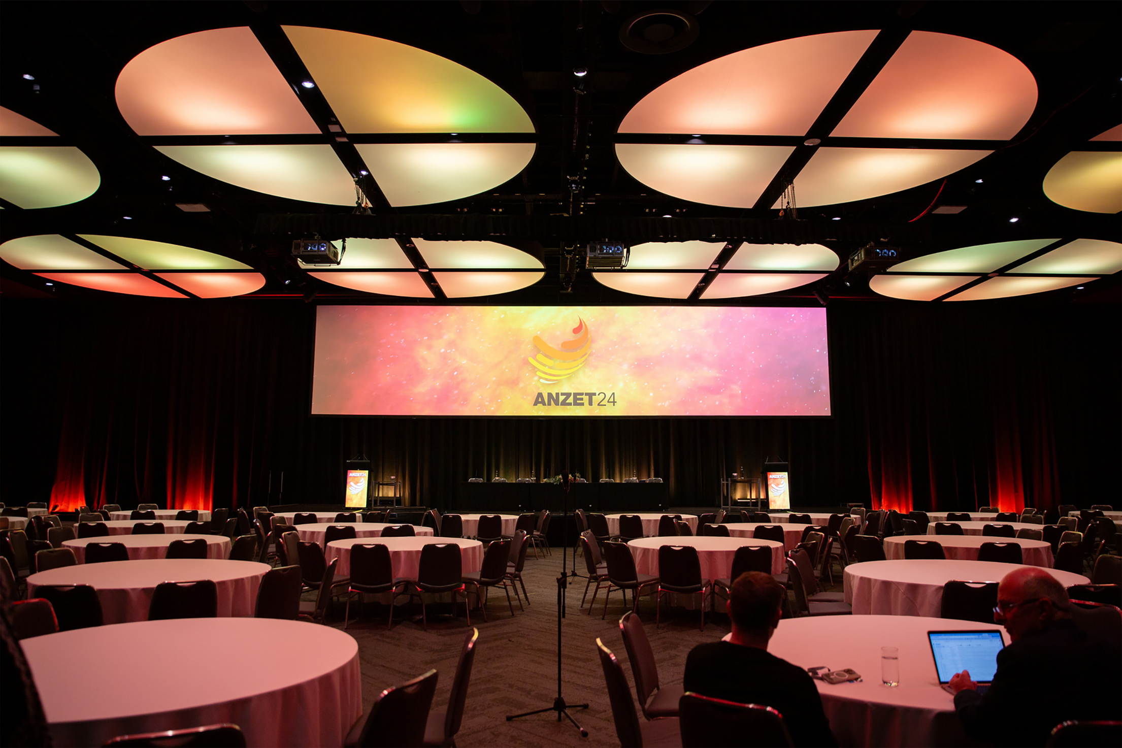 A large conference room with round tables covered with white tablecloths and black chairs, a stage with a large screen displaying 'ANZET24' logo, a black ceiling with illuminated oval panels, and dim lighting.