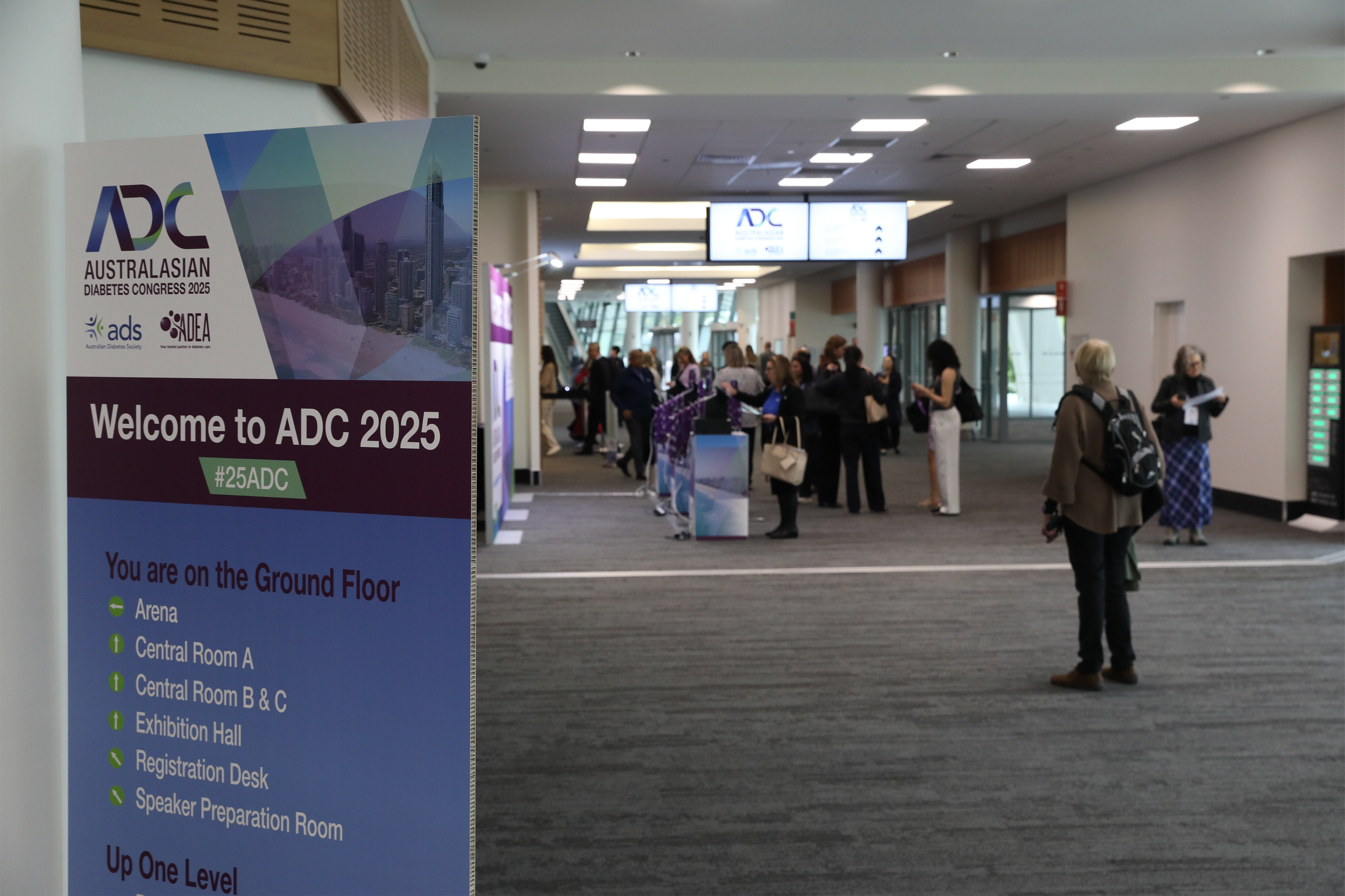 A large indoor conference center with a crowd of people waiting in line and walking. There are two digital screens hanging from the ceiling displaying the ADC logo and information. A purple and white sign in the foreground welcomes attendees to ADC 2025, a diabetes conference, with directions to various rooms and areas on the ground floor.