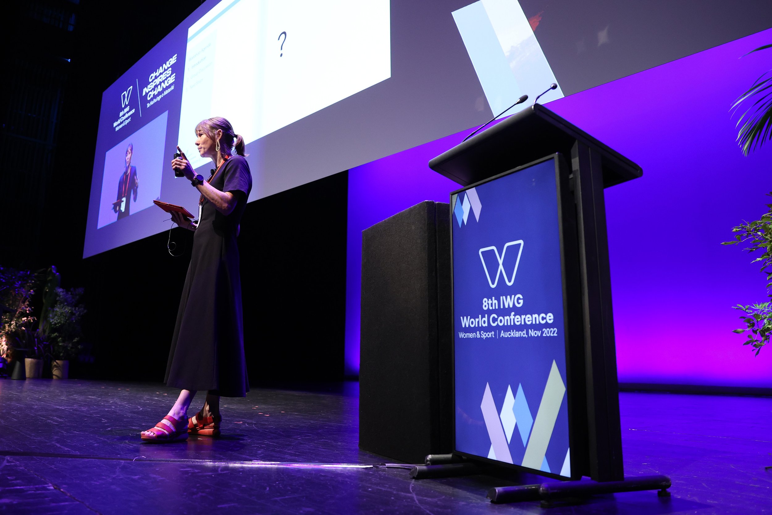 A woman is speaking on stage at the 8th IWG World Conference in Auckland, November 2022. She is holding a microphone and wearing colorful sandals with a black dress. Behind her is a large screen displaying conference information and a podium with the conference logo.