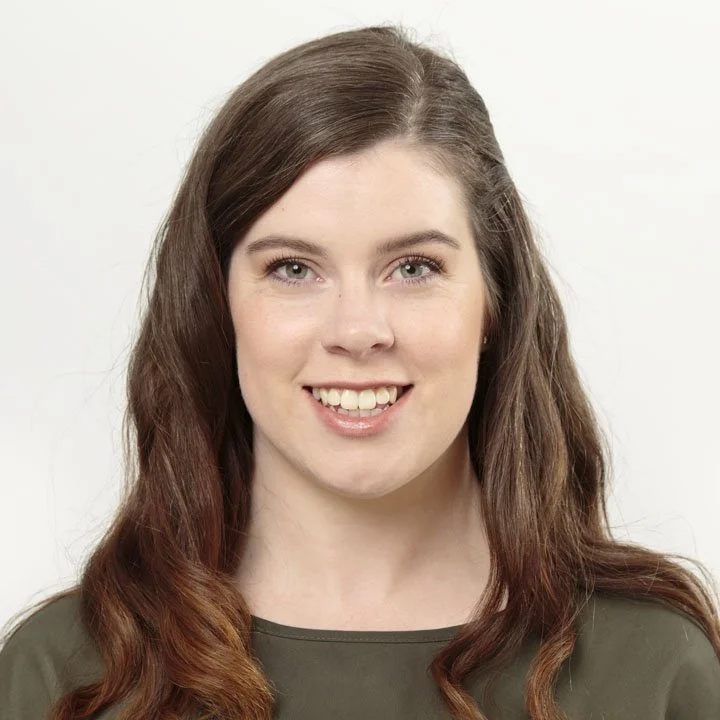 A young woman with long, wavy brown hair and fair skin, smiling in front of a plain white background.
