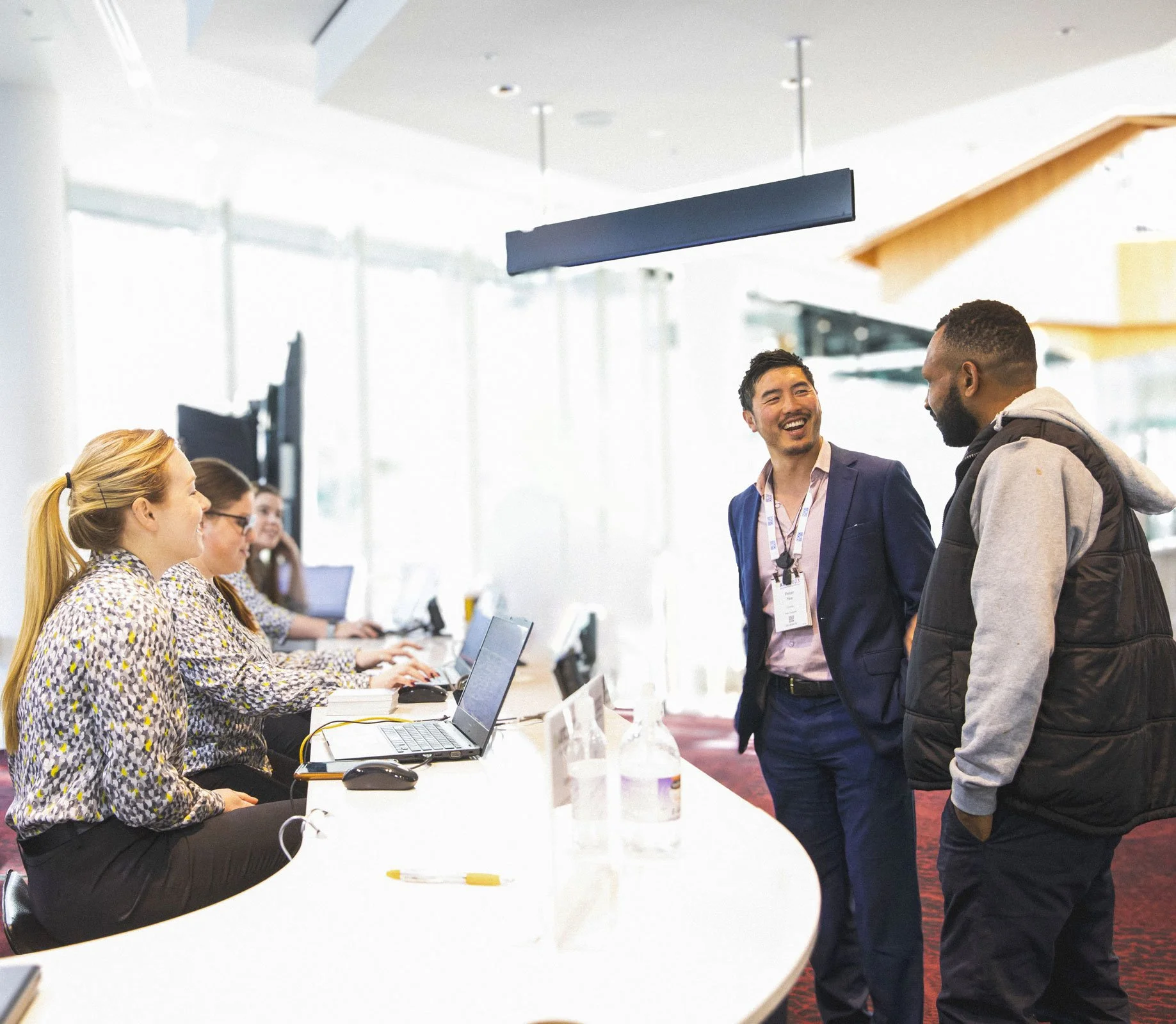 Two women from The Conference Company sitting at a welcome desk at a conference. They are politely greeting two men who are guests at the conference.